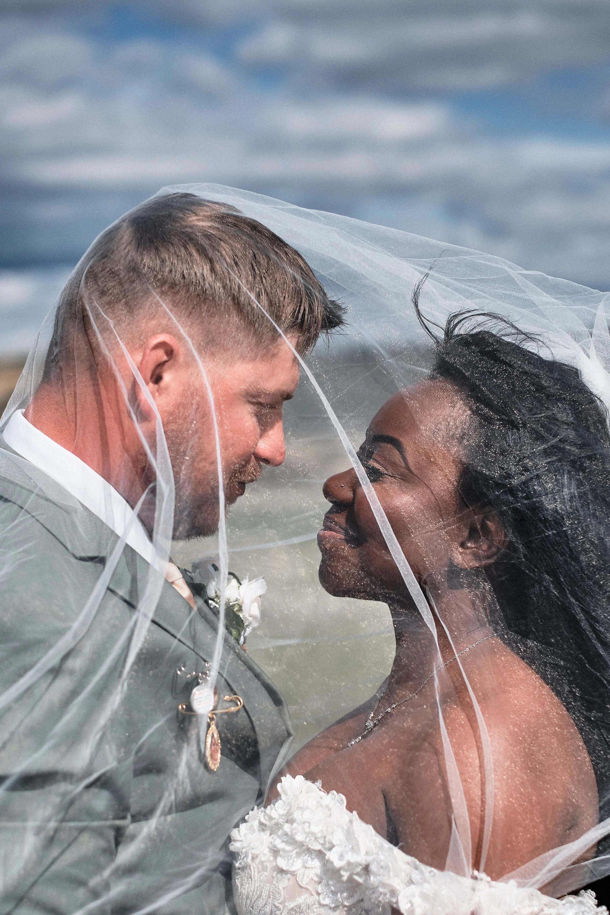 A close-up photograph of a newlywed couple standing close together in the water, with their foreheads touching and eyes closed, both wearing wedding attire and veils, against a background of a cloudy sky at the beach.