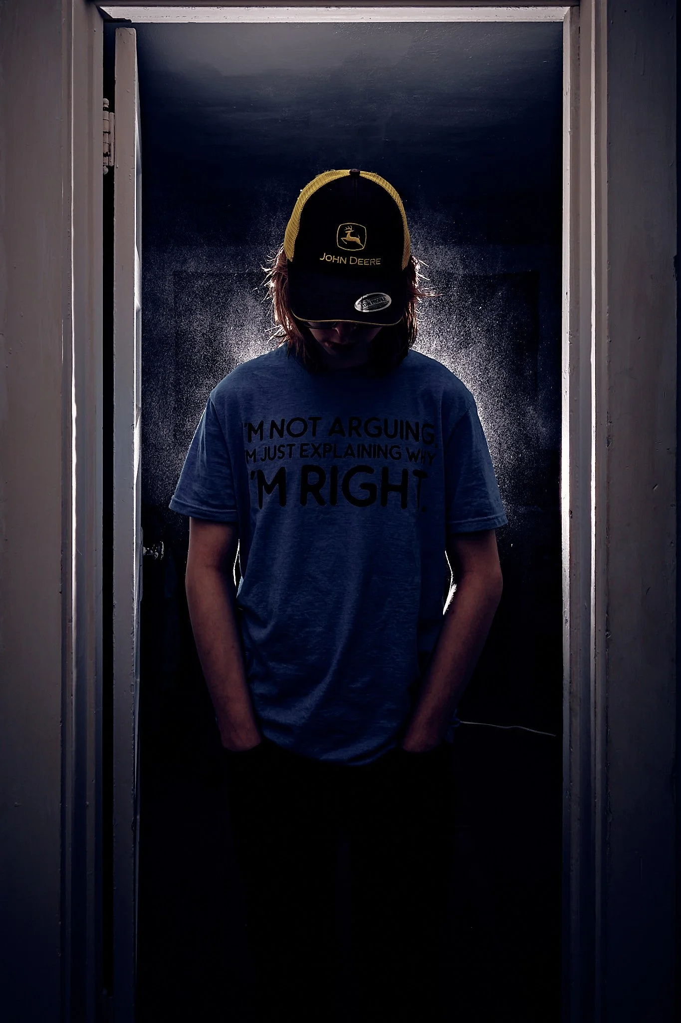 A person standing in a small, dark room or closet with hands in pockets, wearing a John Deere cap and a gray T-shirt that reads 'I'm not arguing, I'm just explaining why I'm right'.