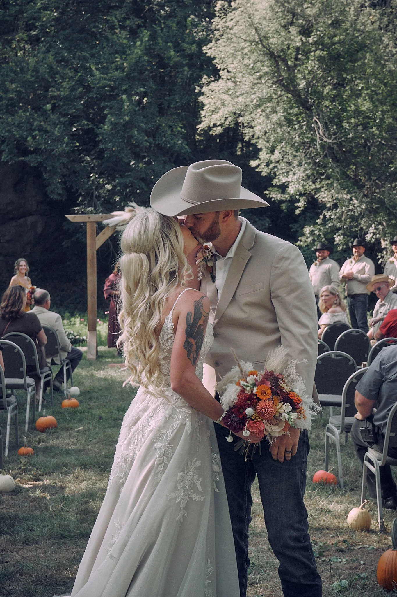 A couple is kissing during their outdoor wedding ceremony, with guests seated in the background and pumpkins decorating the ground.