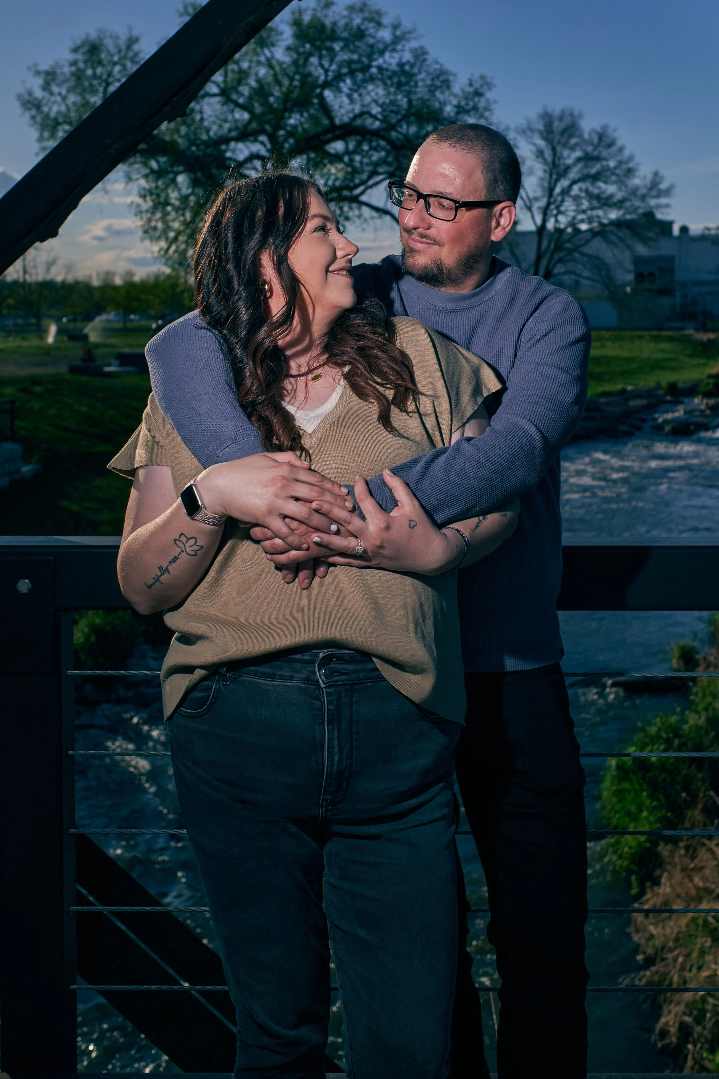 A couple embracing on a bridge outdoors at sunset, smiling at each other with trees and water in the background.