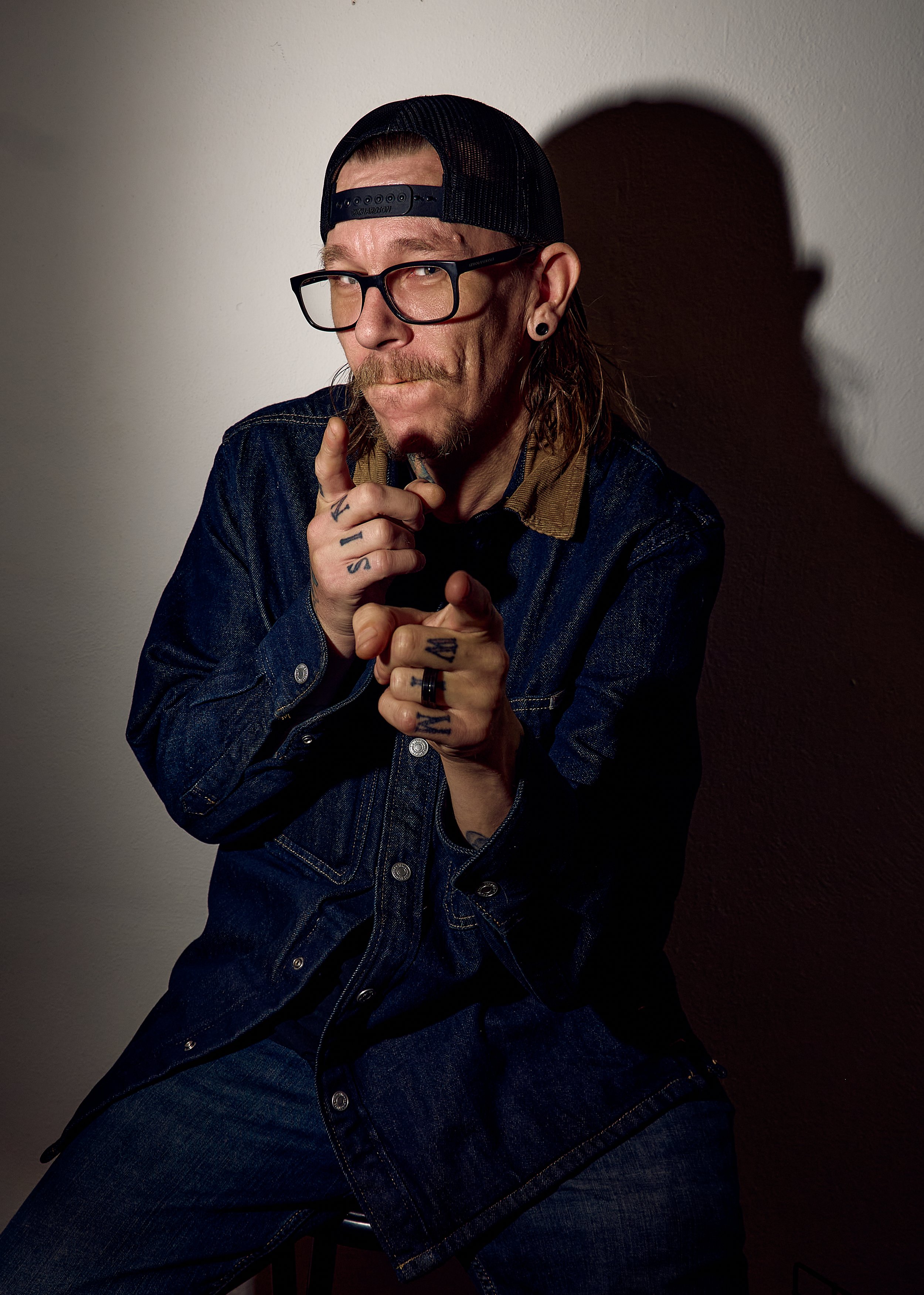 A man with glasses, a baseball cap, tattoos on his fingers, and wearing a denim jacket, sitting and looking at the camera while holding his hands in front of his face against a plain wall.