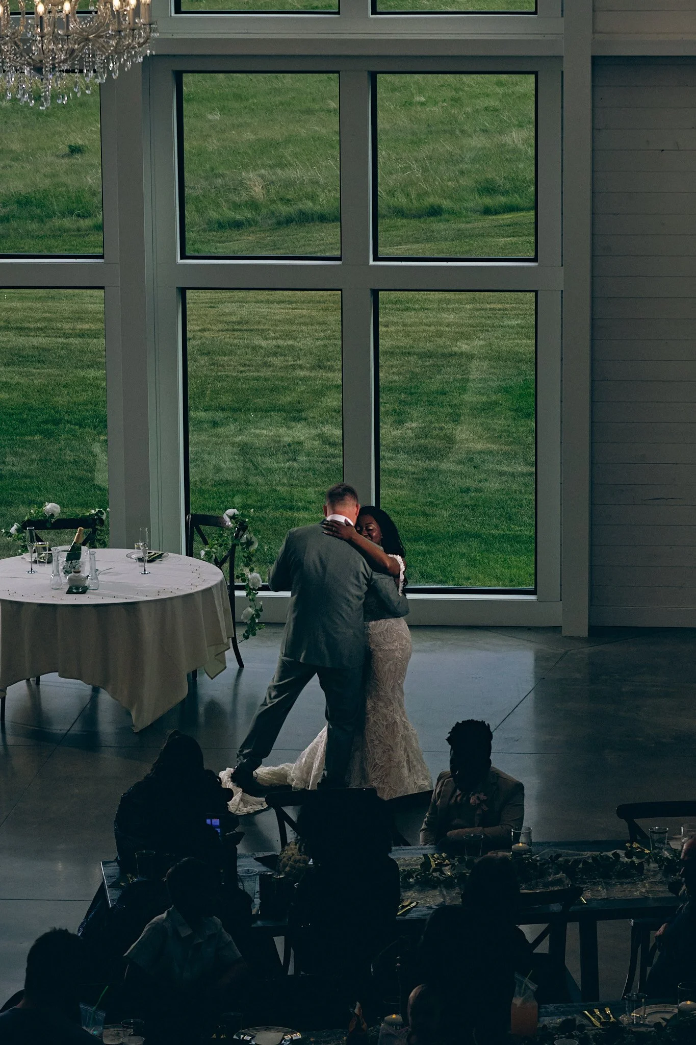 A couple is dancing closely in front of large windows with green grass outside at a wedding reception. There are guest tables with white tablecloths and chairs, and a chandelier hanging from the ceiling.