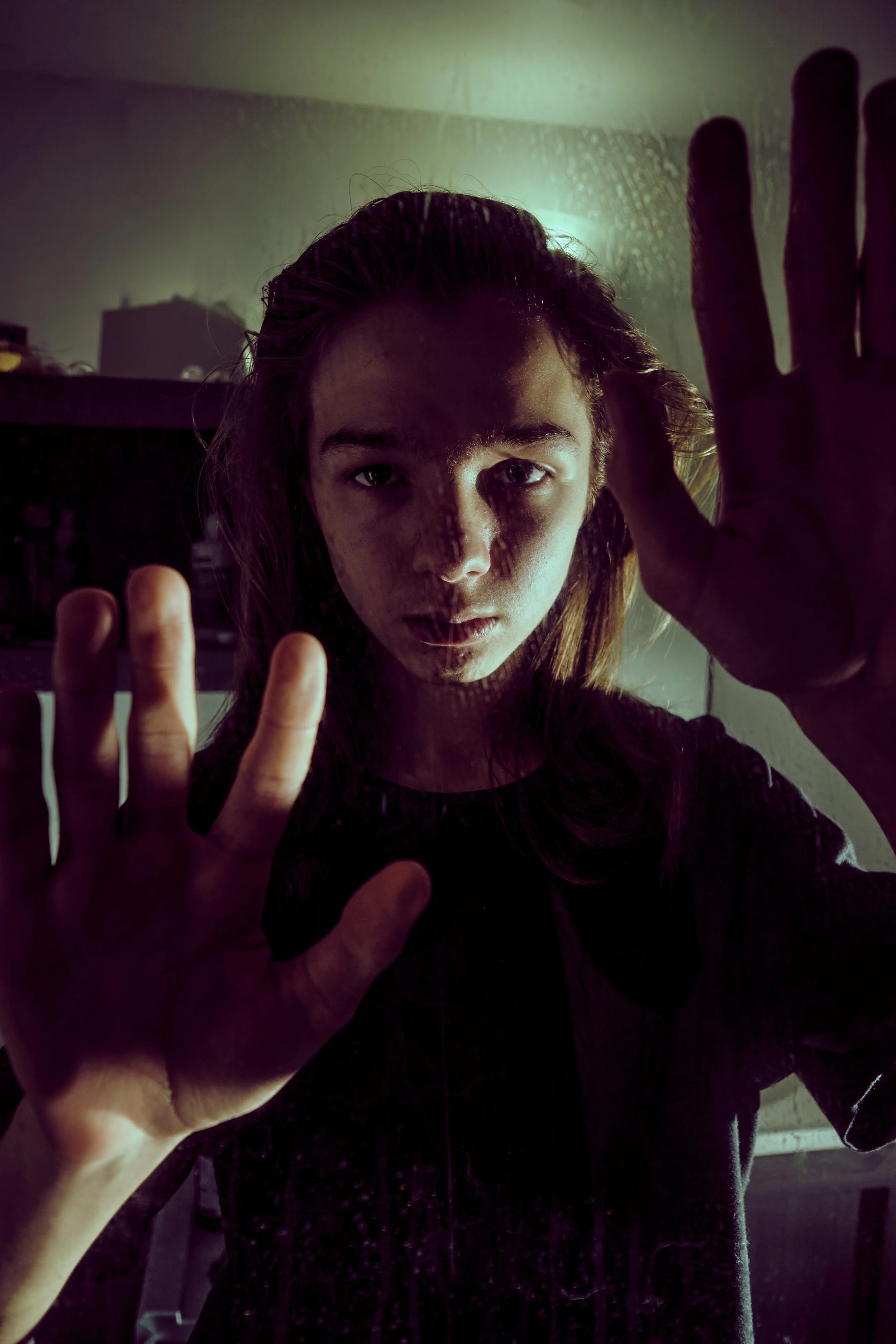 A young man is pressing his face and hands against a glass surface, creating a reflection of her face with a serious expression in a dimly lit room.