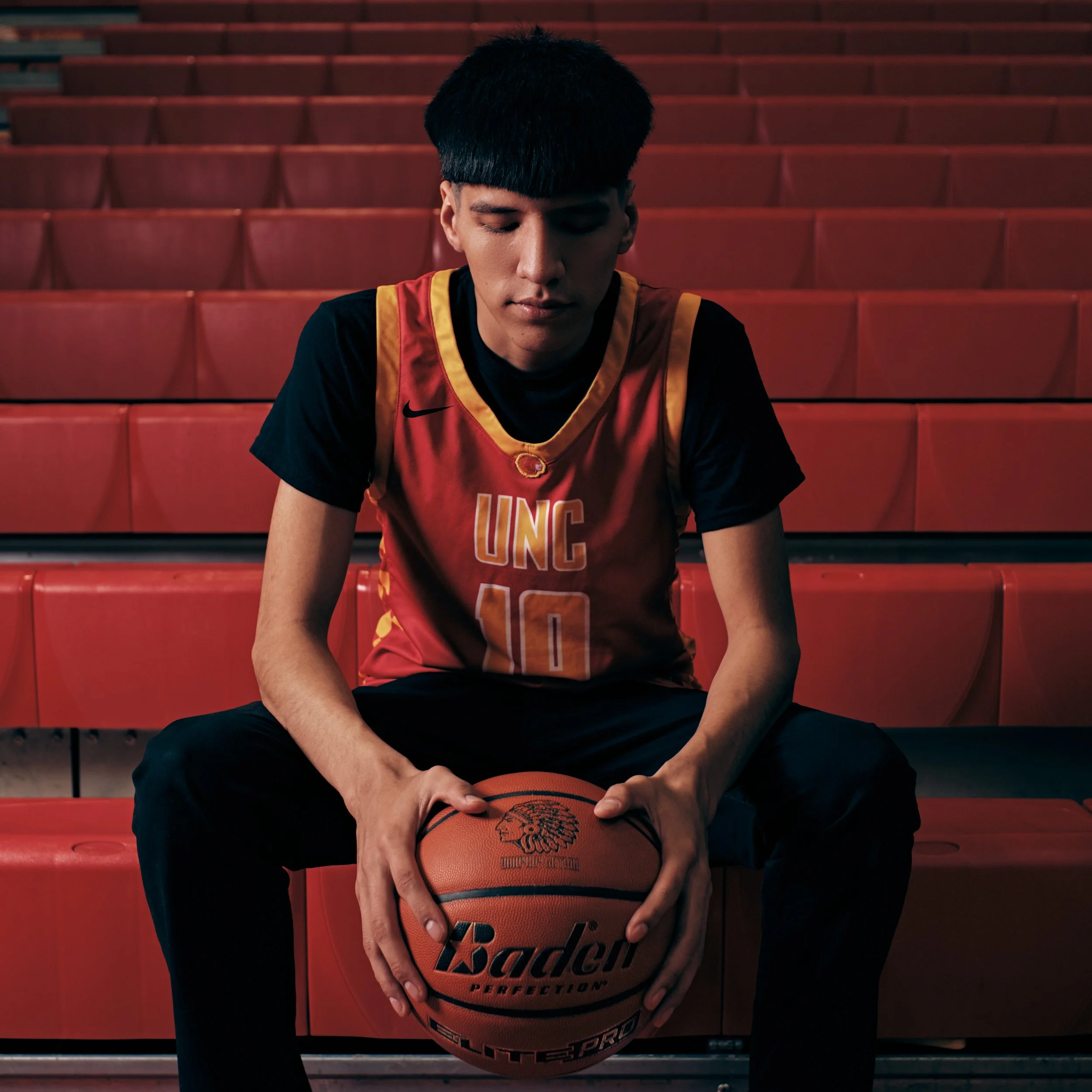A young man with dark hair, wearing a red basketball jersey, seated on red stadium seats, holding a basketball with both hands resting on his lap.