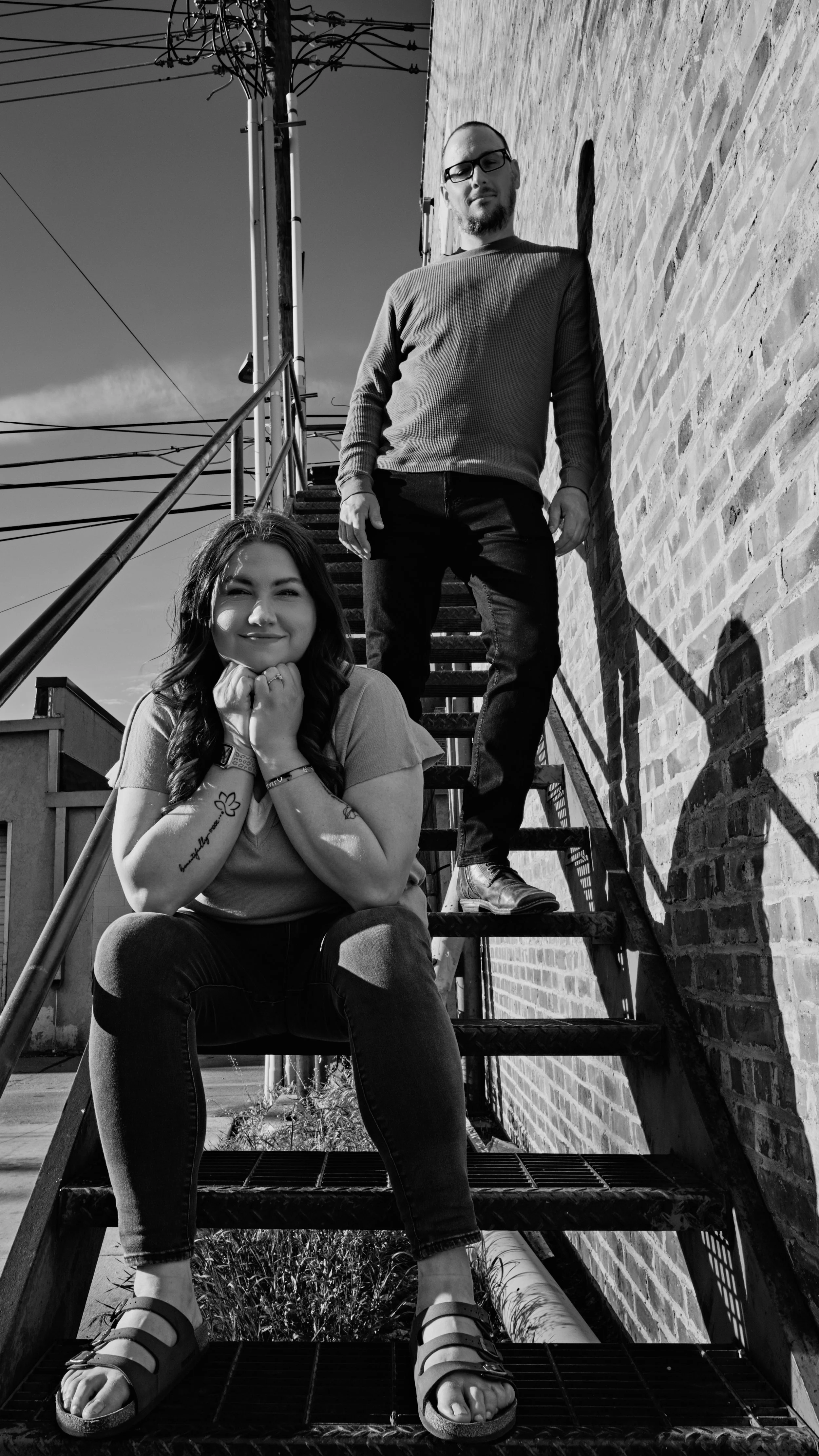 A black and white photo of a young woman sitting on stairs with a young man standing behind her on a metal staircase outside a brick building.