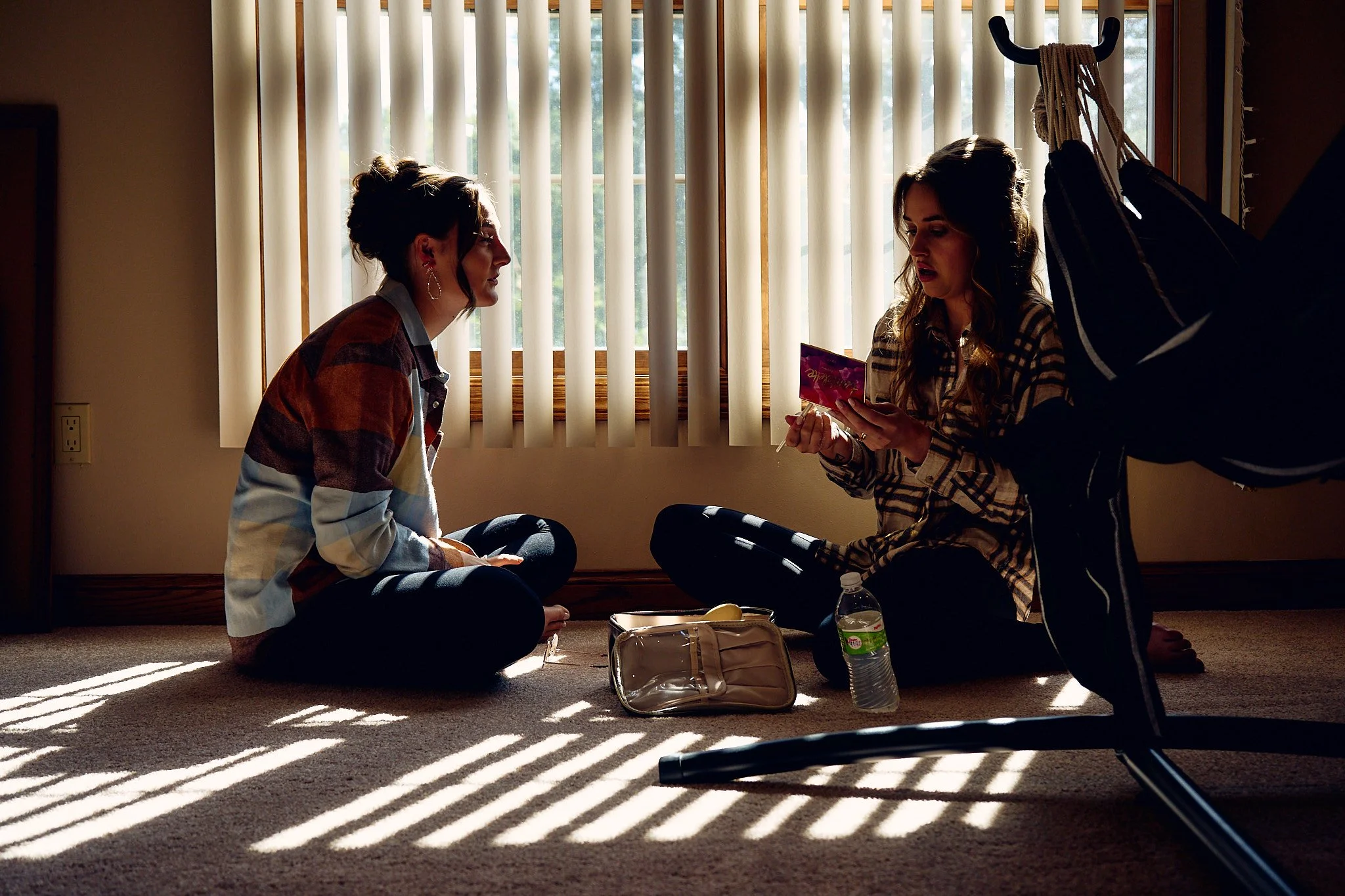 Two women sitting on the floor in front of a window with vertical blinds. One woman is holding a snack packet, and the other is listening. There are personal belongings on the floor, including a water bottle and a small bag.