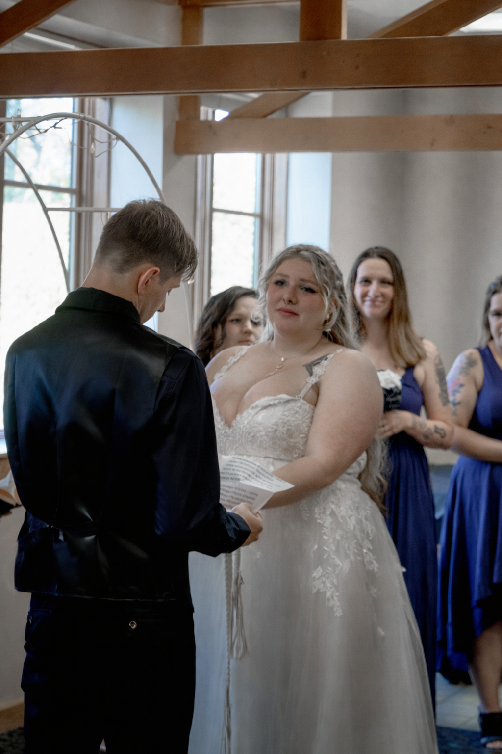 A bride with long blonde hair and a white lace wedding dress looks confused or emotional during a wedding ceremony, reading vows or a speech, while a groom with short hair in a black shirt stands opposite her in front of a group of bridesmaids dresse