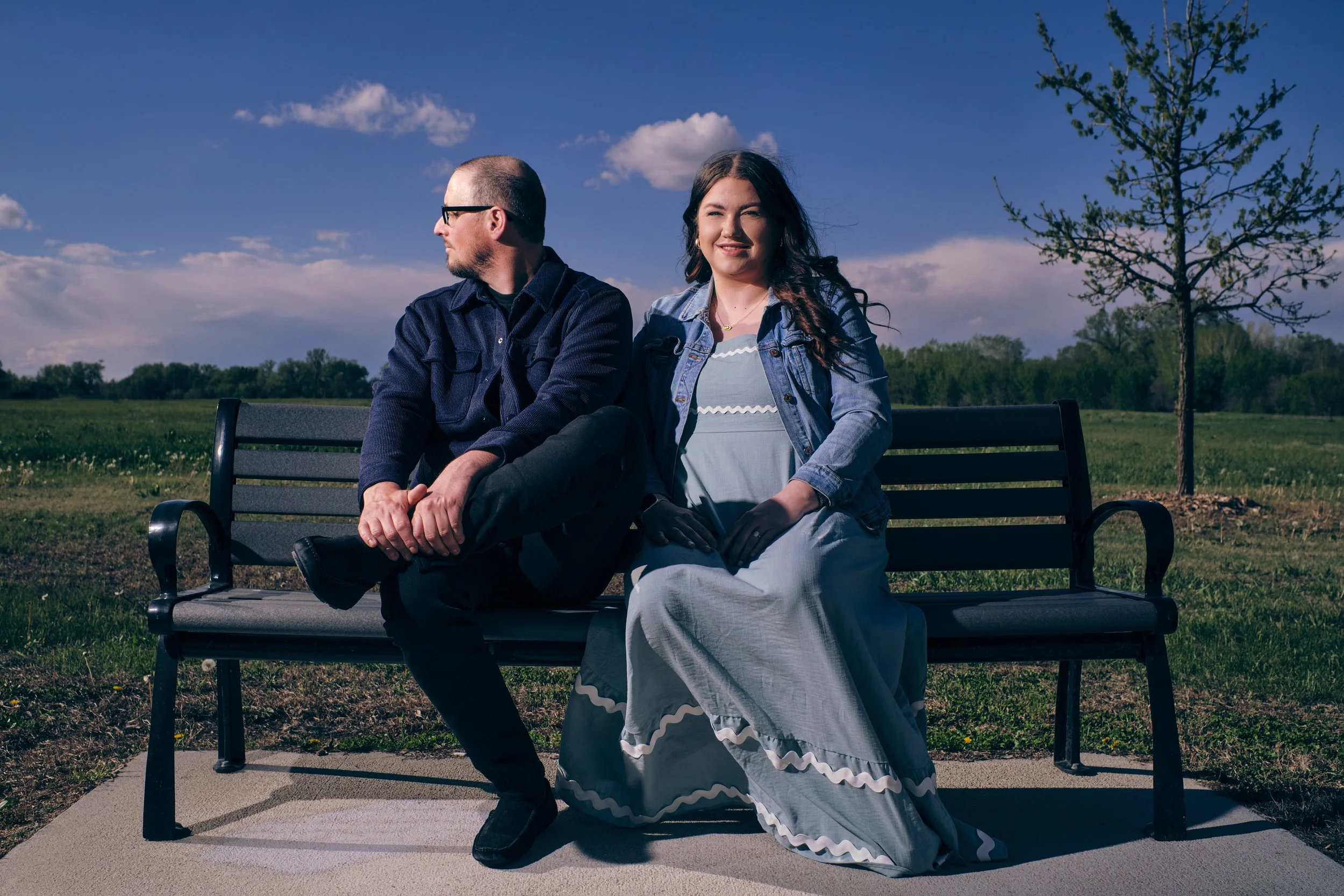 A man and woman sitting on a park bench outdoors on a sunny day, with green grass and trees in the background, and a partly cloudy sky.