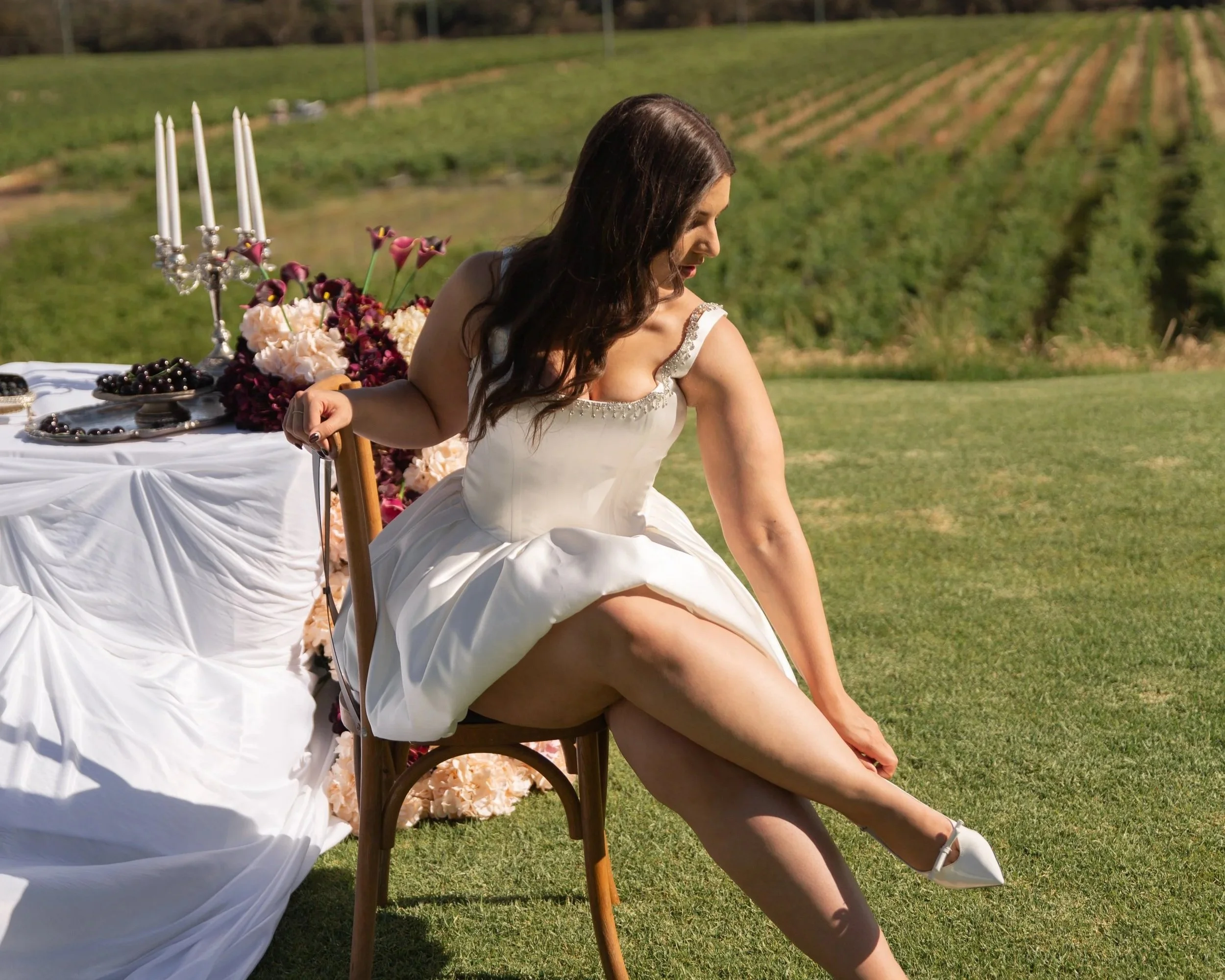 A woman in a wedding dress sitting on a chair outdoors on a grassy field, with a decorated table and scenic vineyard in the background.