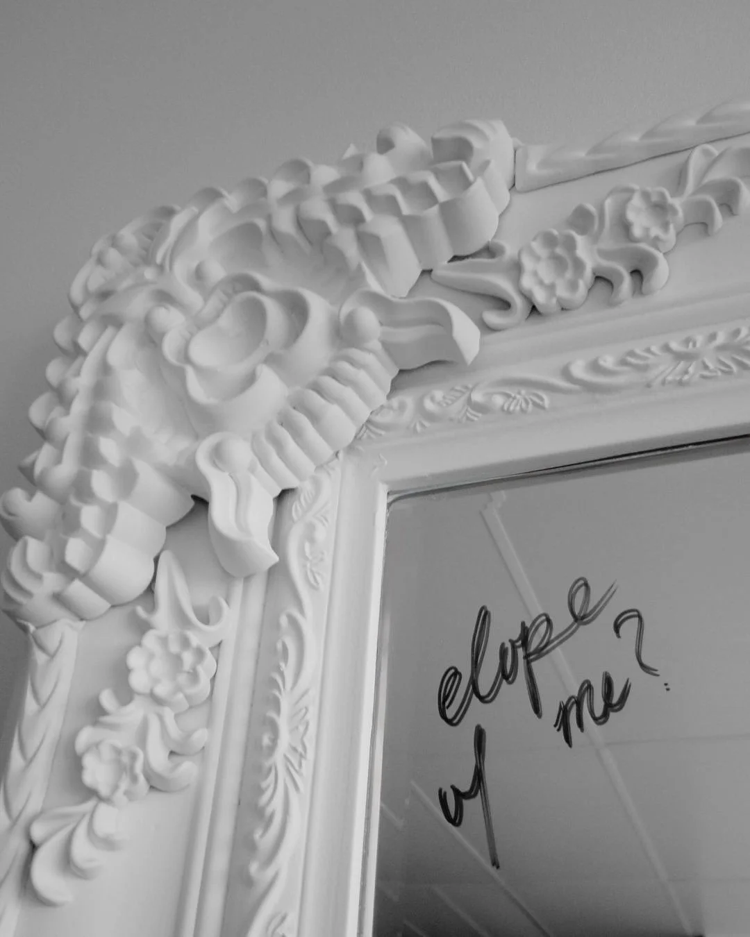 Decorative ornate mirror frame with intricate floral and scroll carvings, reflecting a wall with a handwritten note that says 'elope w me?' in black ink.