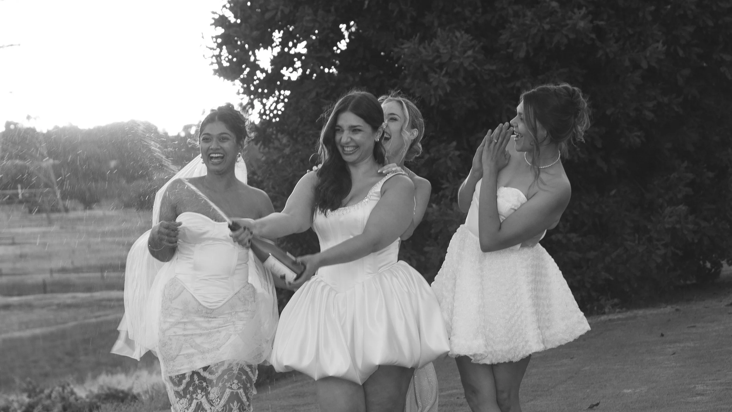 Four women in wedding dresses celebrating outdoors with champagne, laughing and smiling.