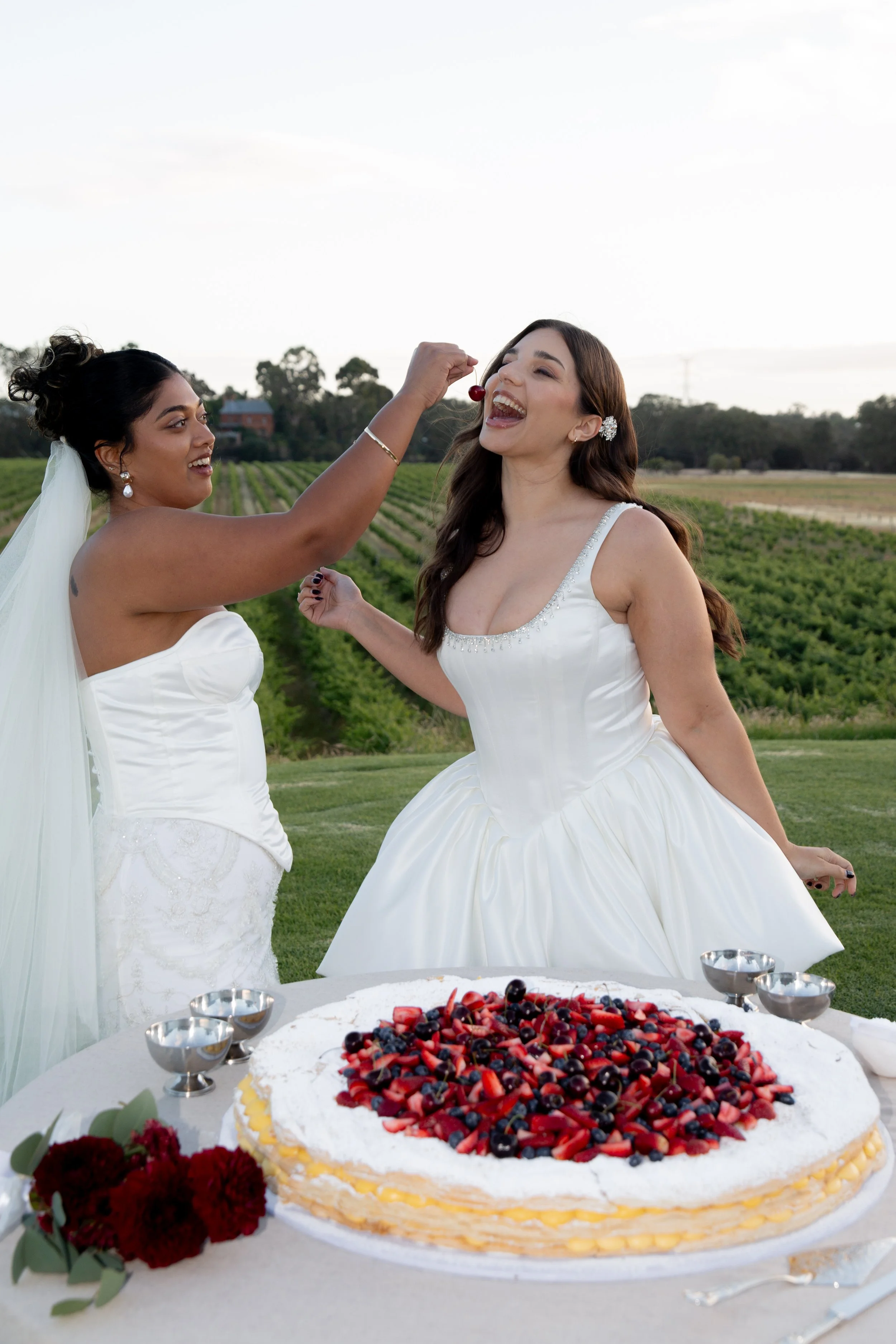Two women in wedding dresses celebrating outdoors near a large berry-topped cake, with one feeding cherries to the other, and a scenic green landscape in the background.