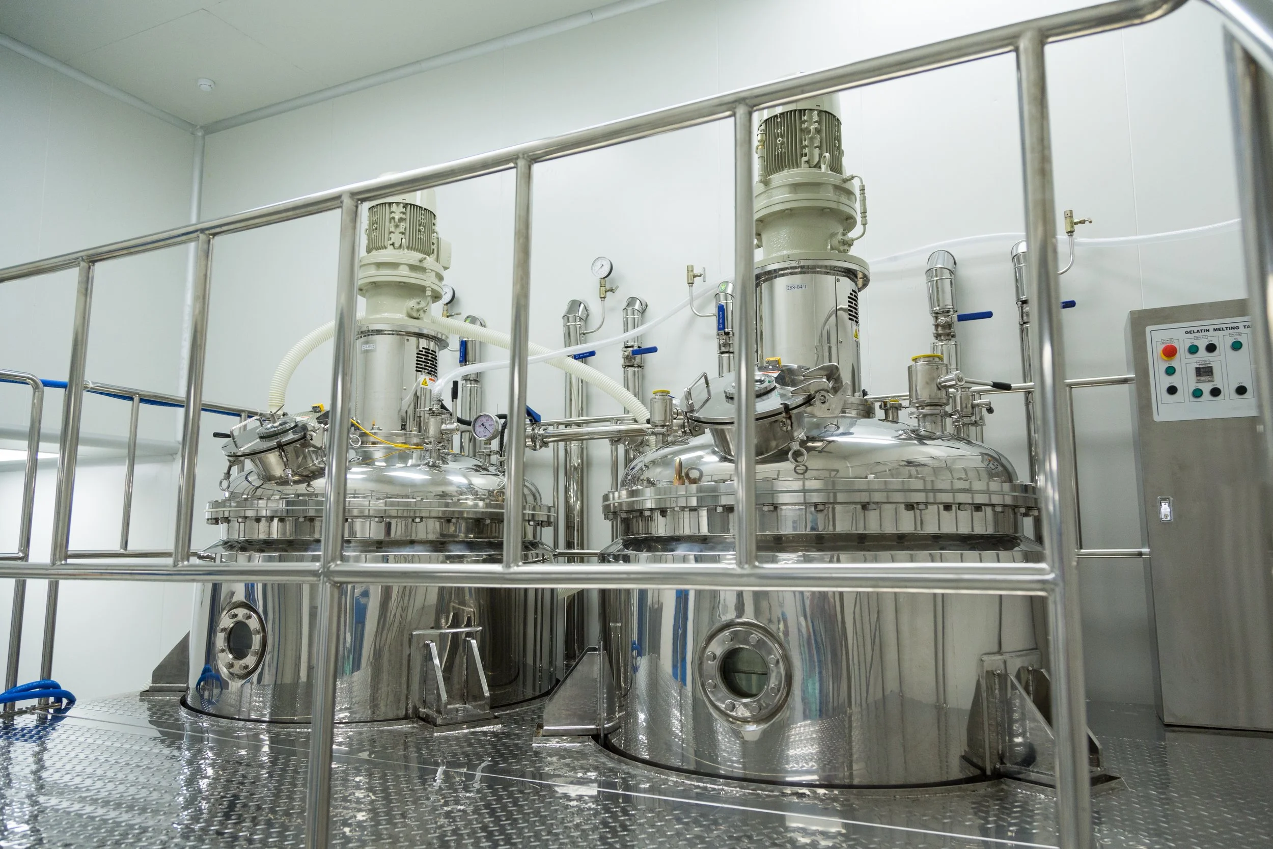 Industrial stainless steel equipment with large tanks, pipes, gauges, and control panel in a clean, white laboratory or manufacturing facility.