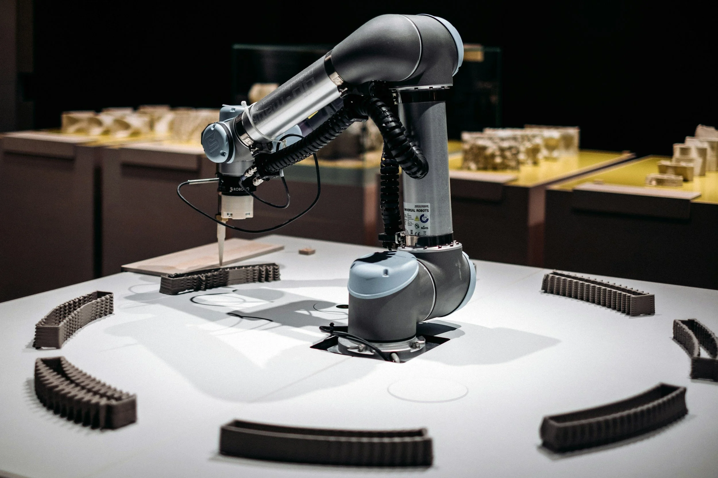 A robotic arm with a pen attached, writing on a small surface, surrounded by brown curved track pieces on a white table.