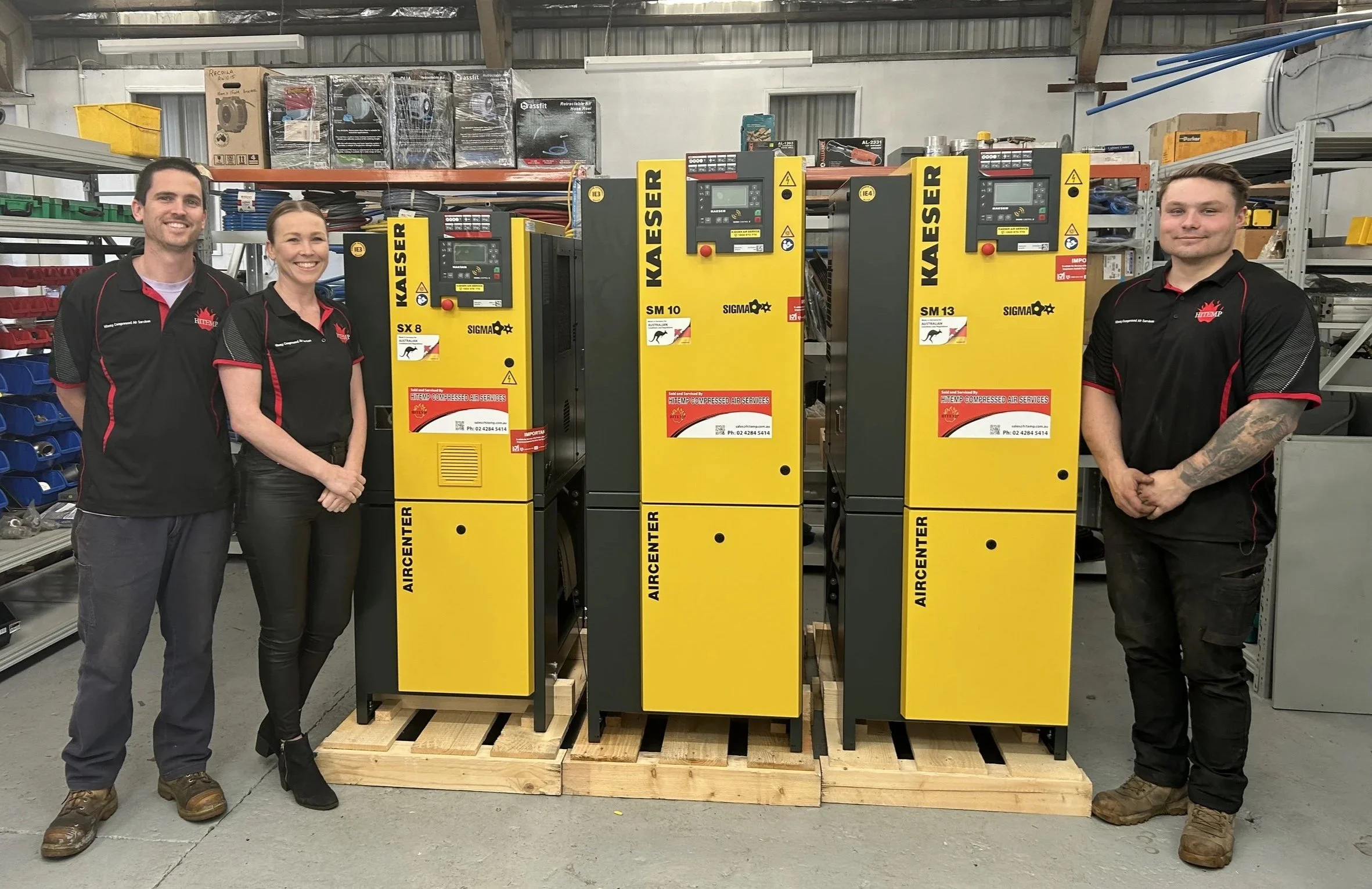 Four people standing beside yellow and black compressed air systems in a warehouse, with shelves and boxes in the background.