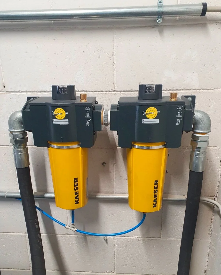 Two paired industrial water filtration units with yellow filters labeled 'KAESER' mounted on a wall, connected by pipes and a blue tube.