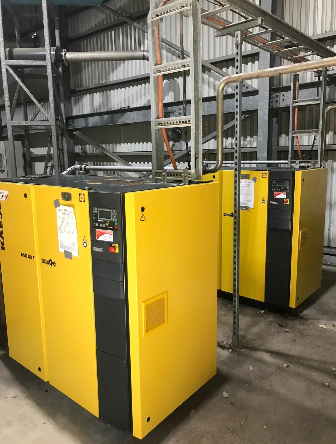 Two yellow industrial chillers in a warehouse setting with metal shelving and pipes.