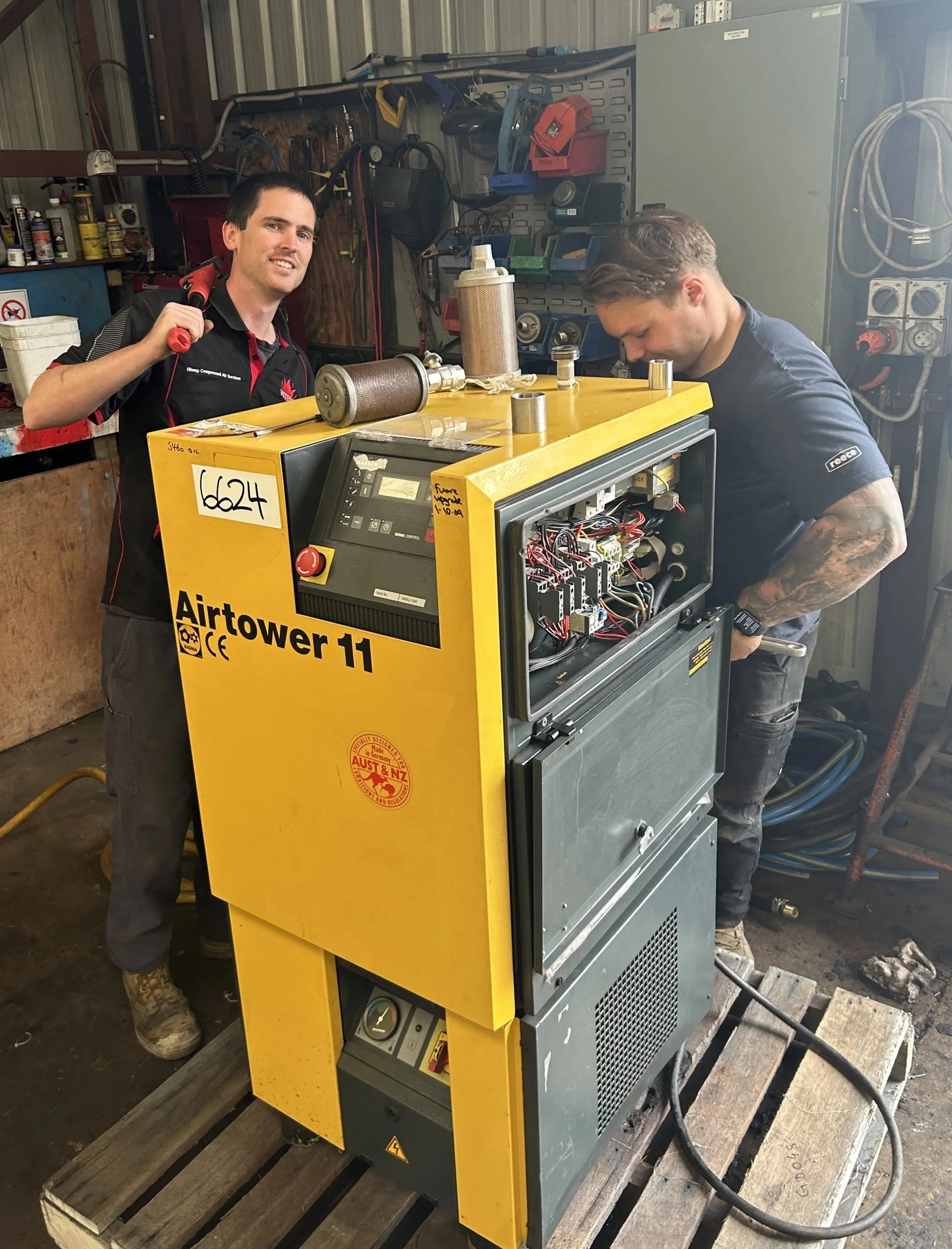 Two men working on a yellow air conditioning or refrigeration unit, with various tools and equipment around in a workshop.