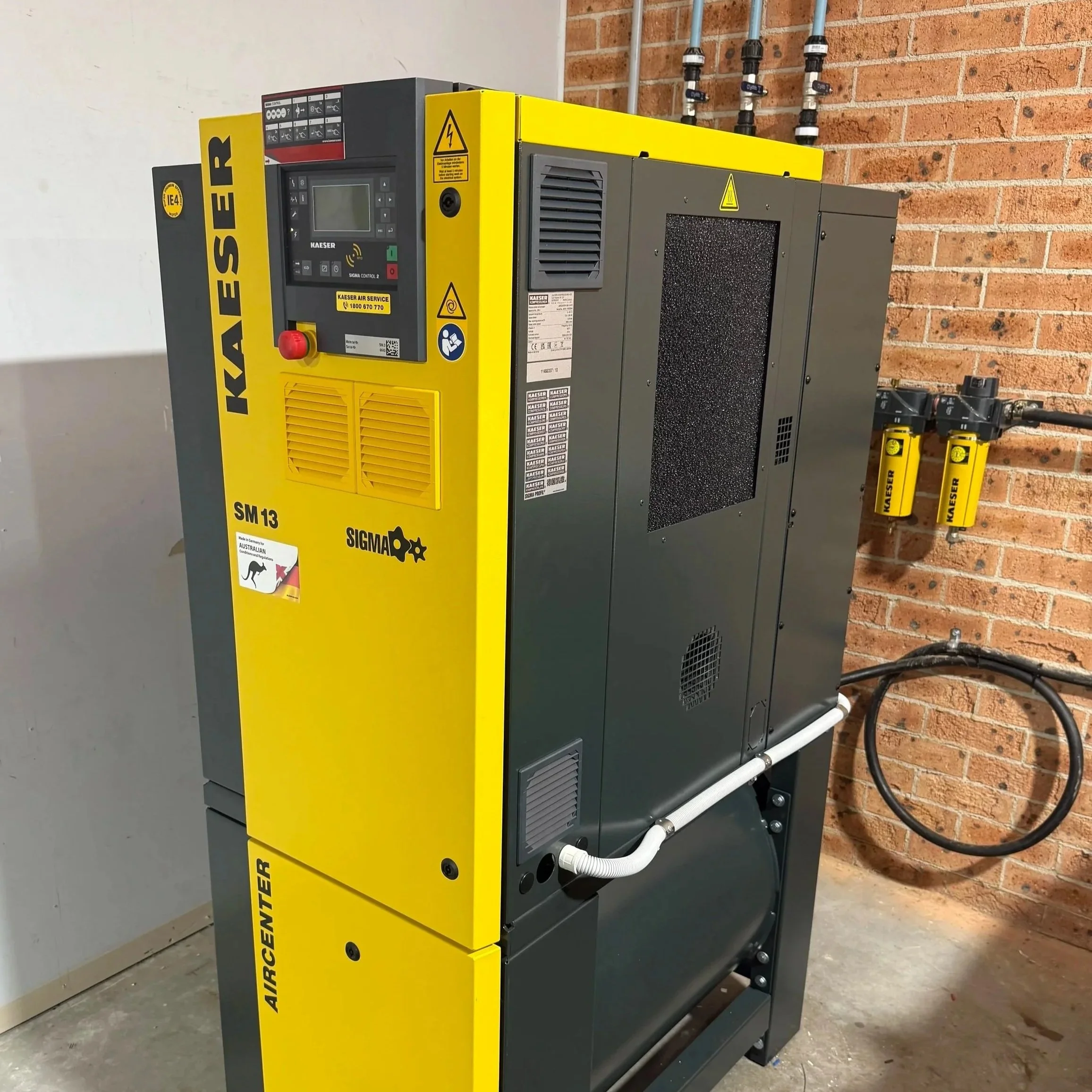 A yellow and gray industrial air compressor unit with control panel, warning labels, and connections on a concrete floor against a brick wall.