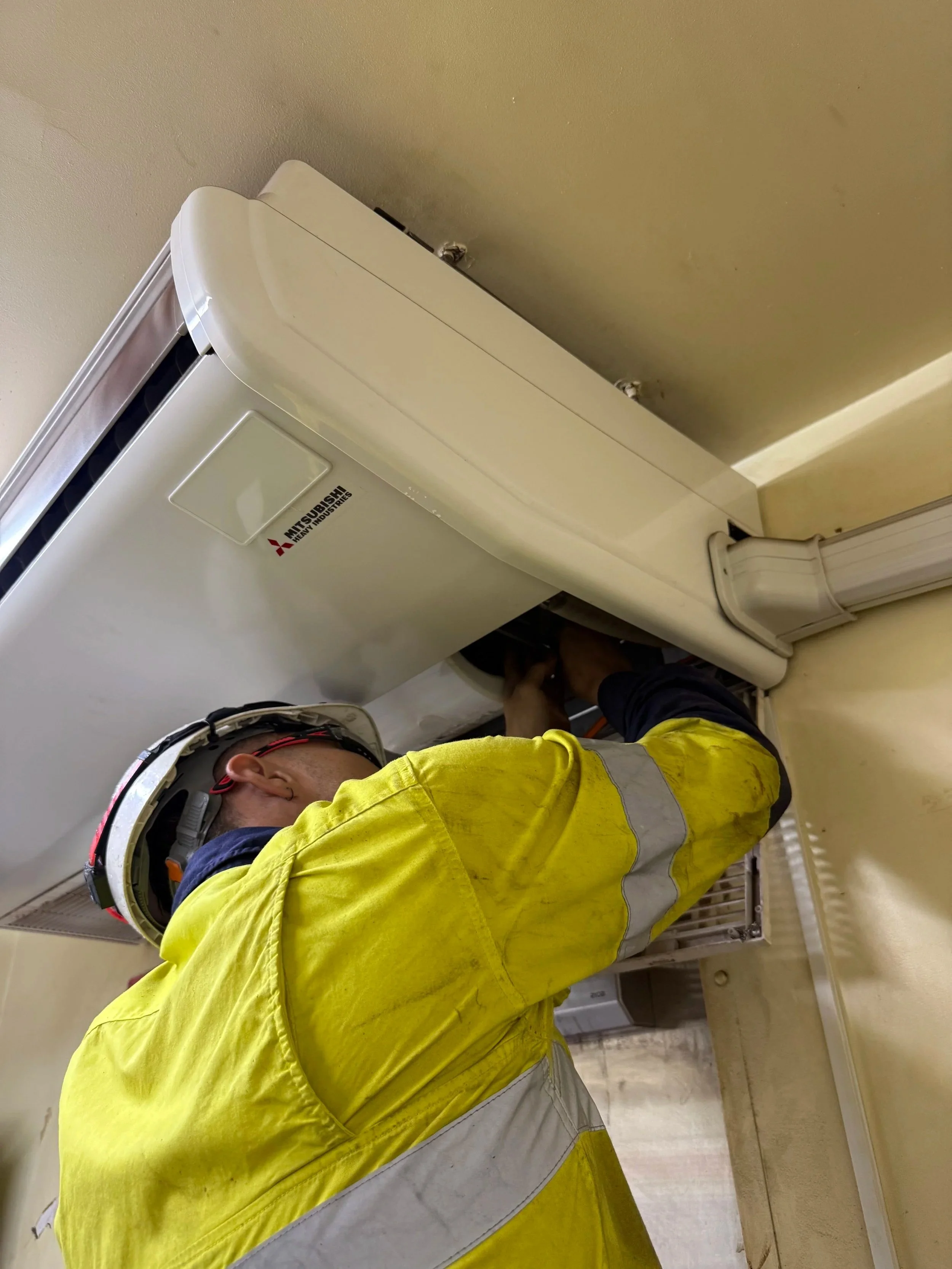 A technician wearing a yellow safety jacket and a helmet working on an air conditioning unit.