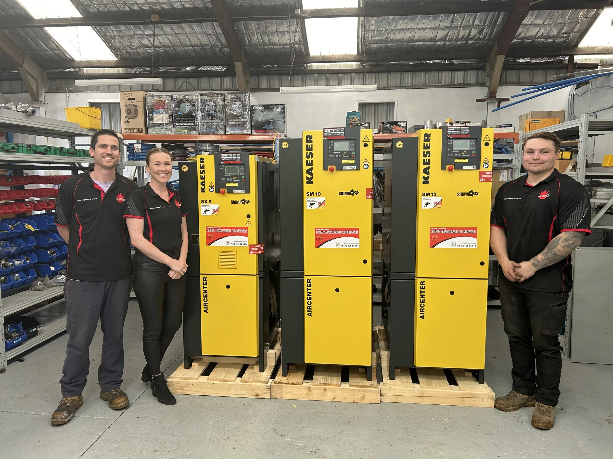 Three people standing next to three yellow industrial machines labeled Kaiser, in a workshop or warehouse setting.
