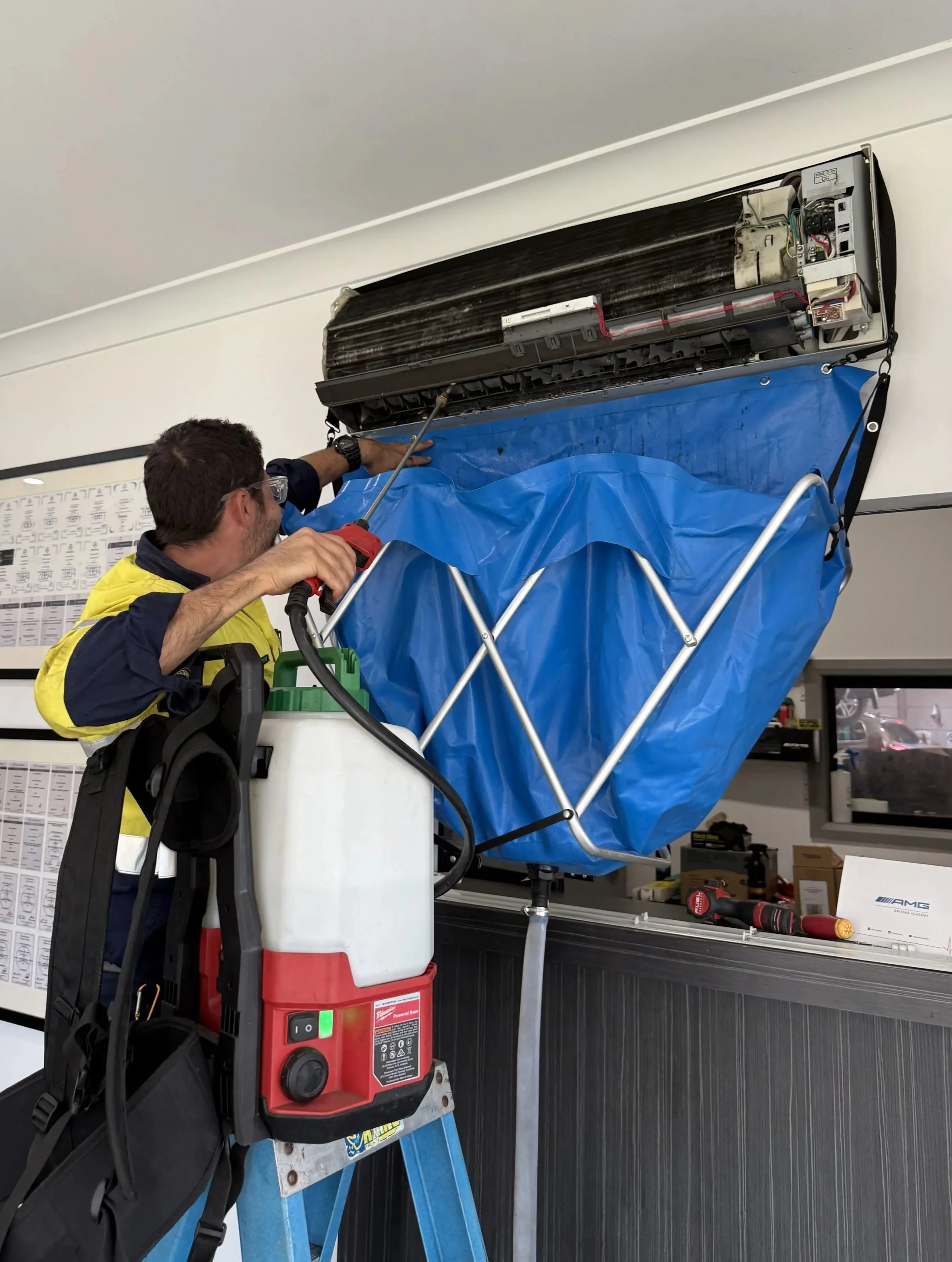 Technician repairing wall-mounted air conditioning unit with tools and a blue protective cover, using a ladder inside a room.