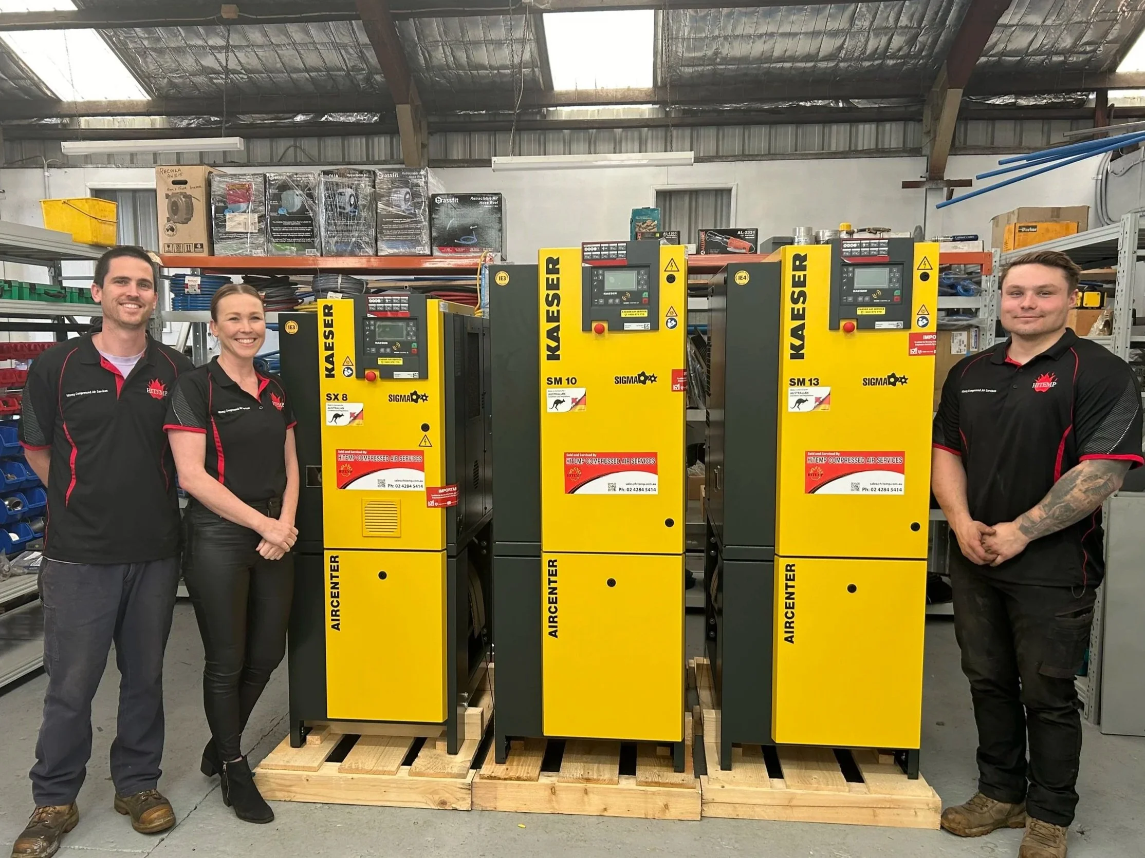 Four people standing in front of yellow industrial air compressor units inside a warehouse, shelves with equipment and boxes in the background.