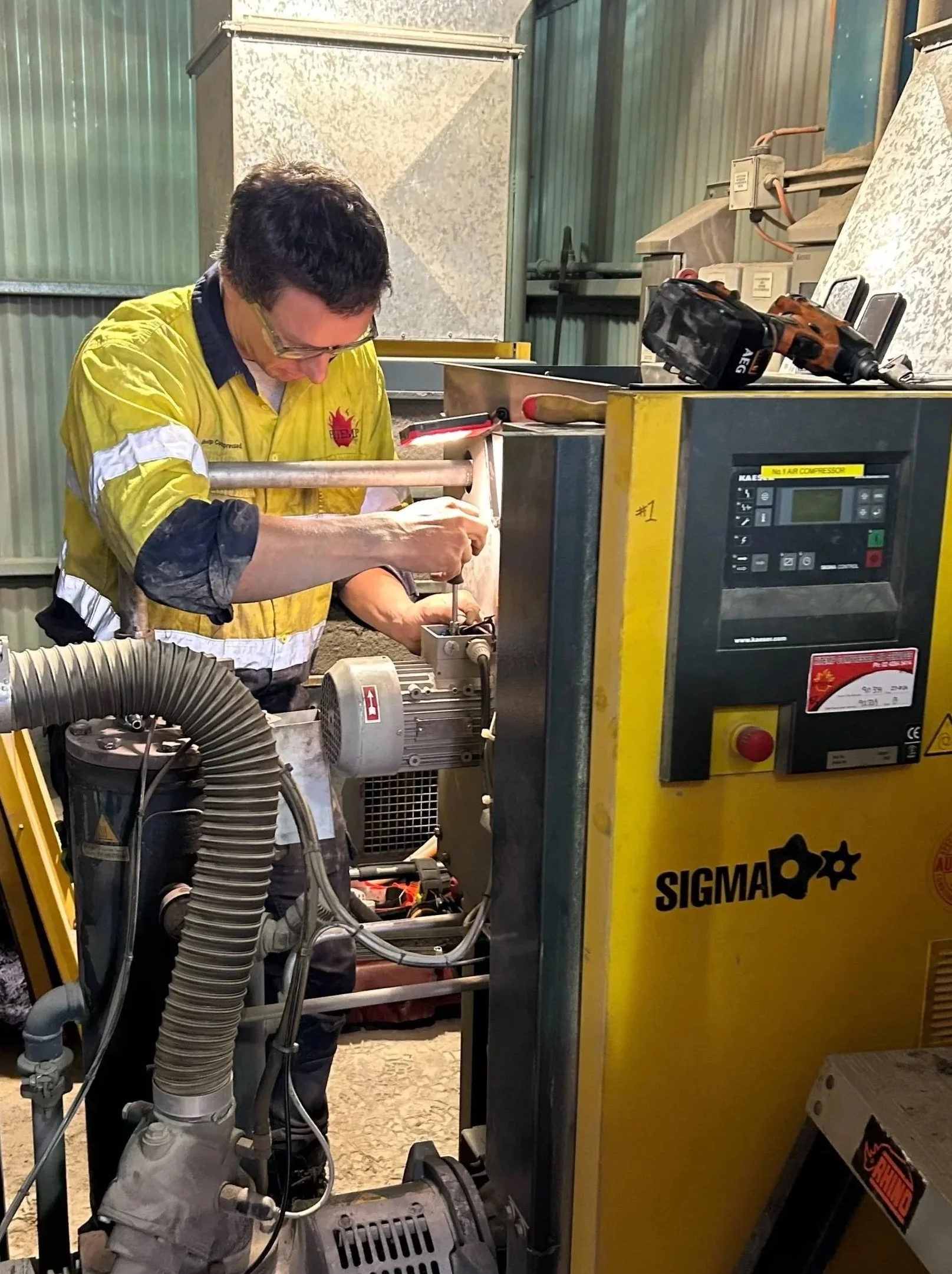 A man wearing safety glasses and a yellow work shirt is working on a large industrial machine labeled 'SIGMA'. The man is focused on the machine, and various tools and a drill are placed on top of the machine. The setting appears to be an industrial or workshop environment with metallic walls and equipment.