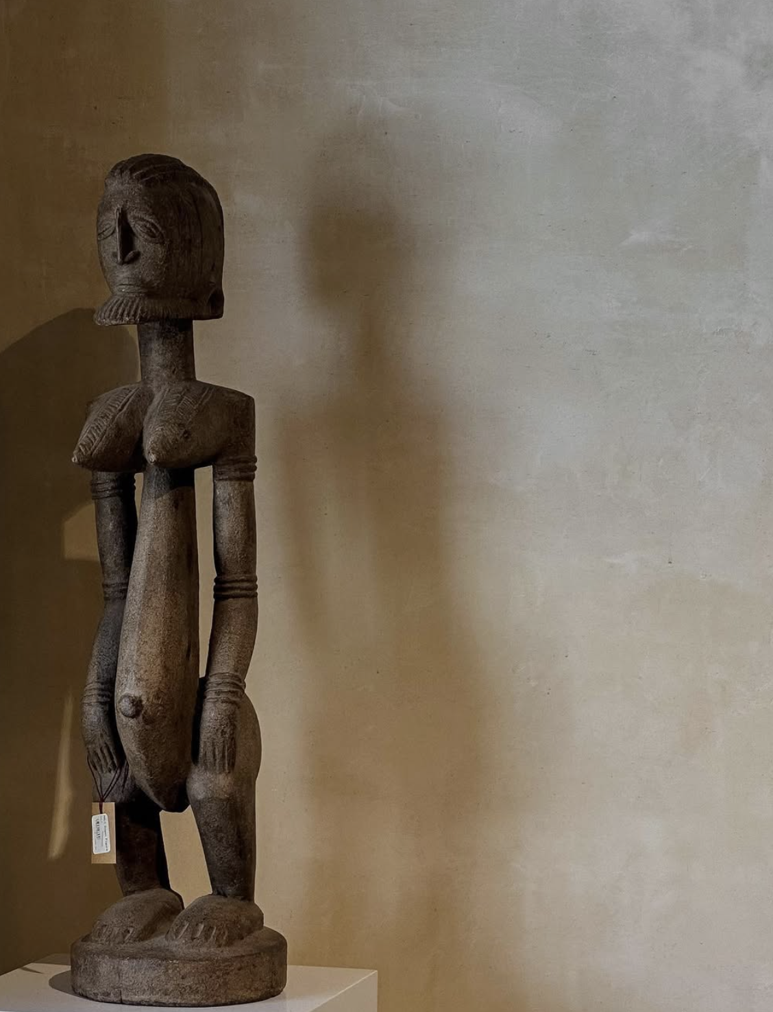 African tribal wooden sculpture of a standing human figure with elongated limbs, detailed facial features, and a rounded head, displayed on a white pedestal against a neutral wall.