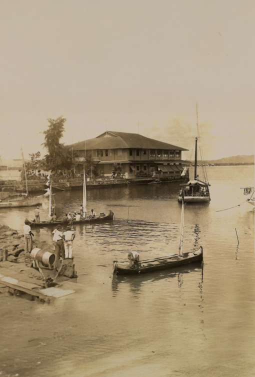 Vintage photo of boats docked by a riverside building with people on the shore, in sepia tone.