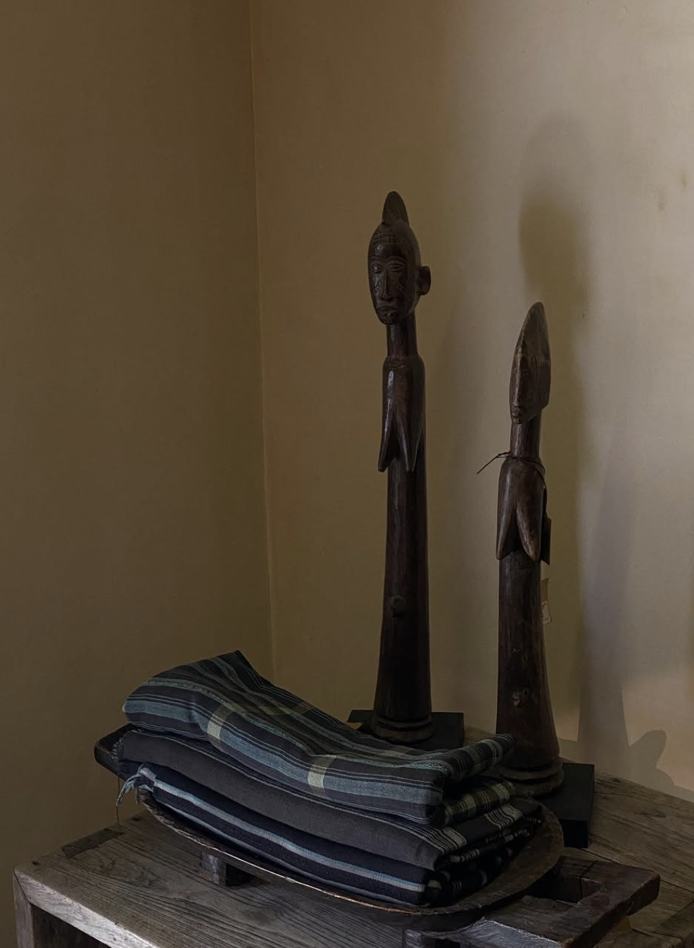 Two wooden African tribal statues on a black stand placed on a rustic wooden surface, with rolled fabric on a tray in front of them, against a beige wall.