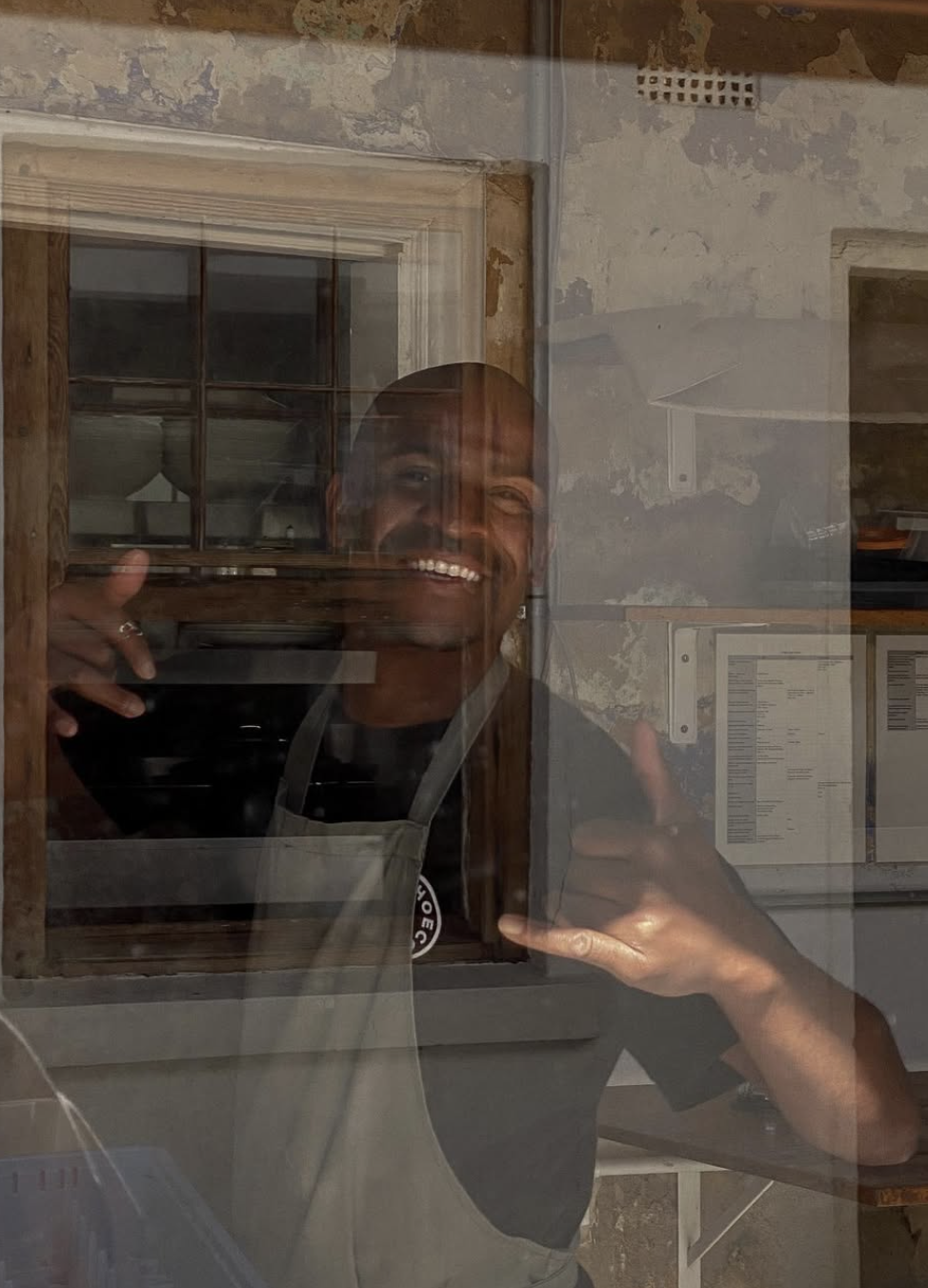 Person smiling and giving a thumbs-up behind a glass window, with a rustic interior in the background.