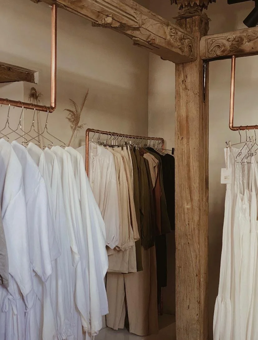 Clothing store with white, beige, brown, and black garments on racks, rustic wooden beams and posts, and copper pipe shelving.
