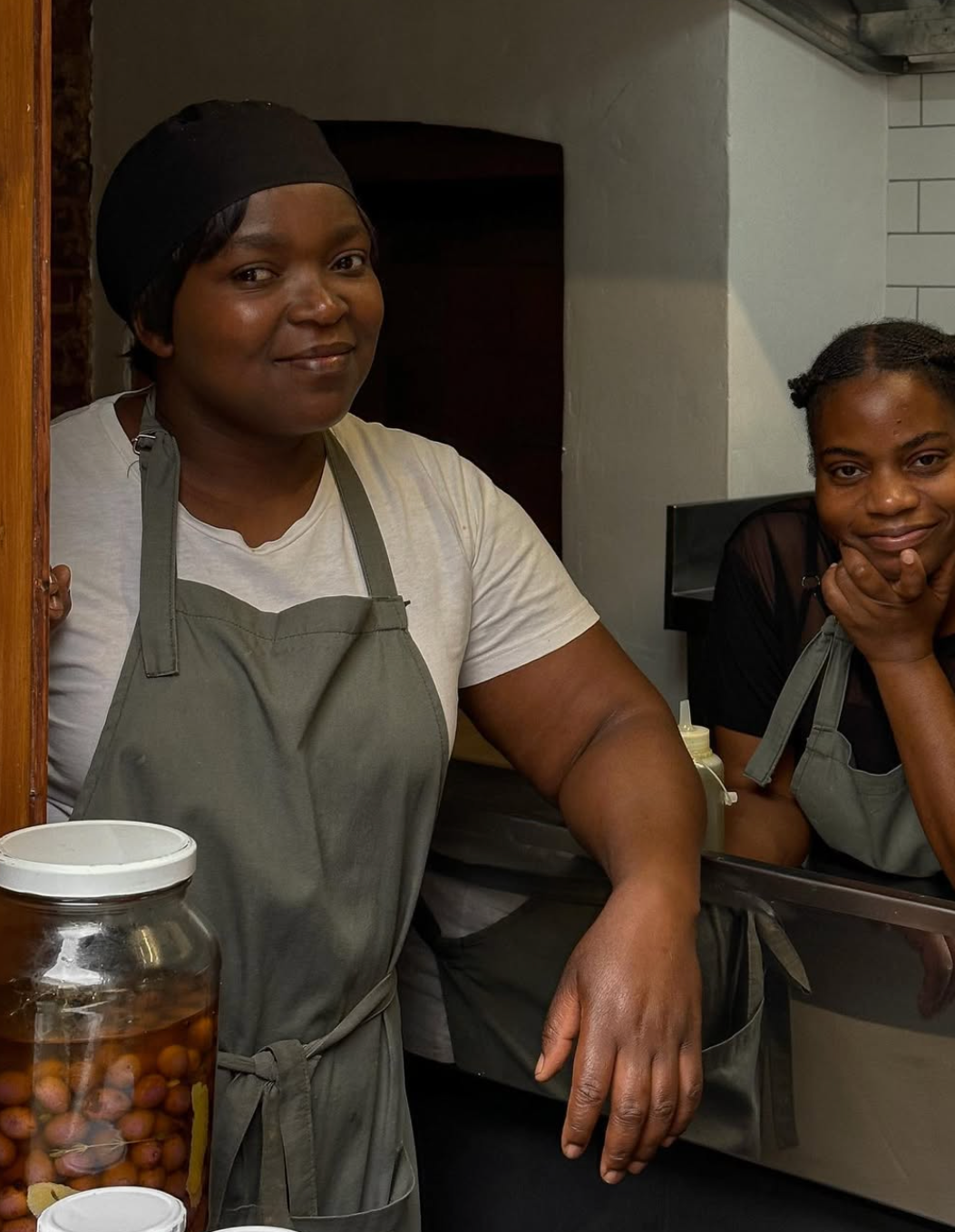 Two women wearing aprons, standing behind a counter, smiling at the camera in a kitchen.