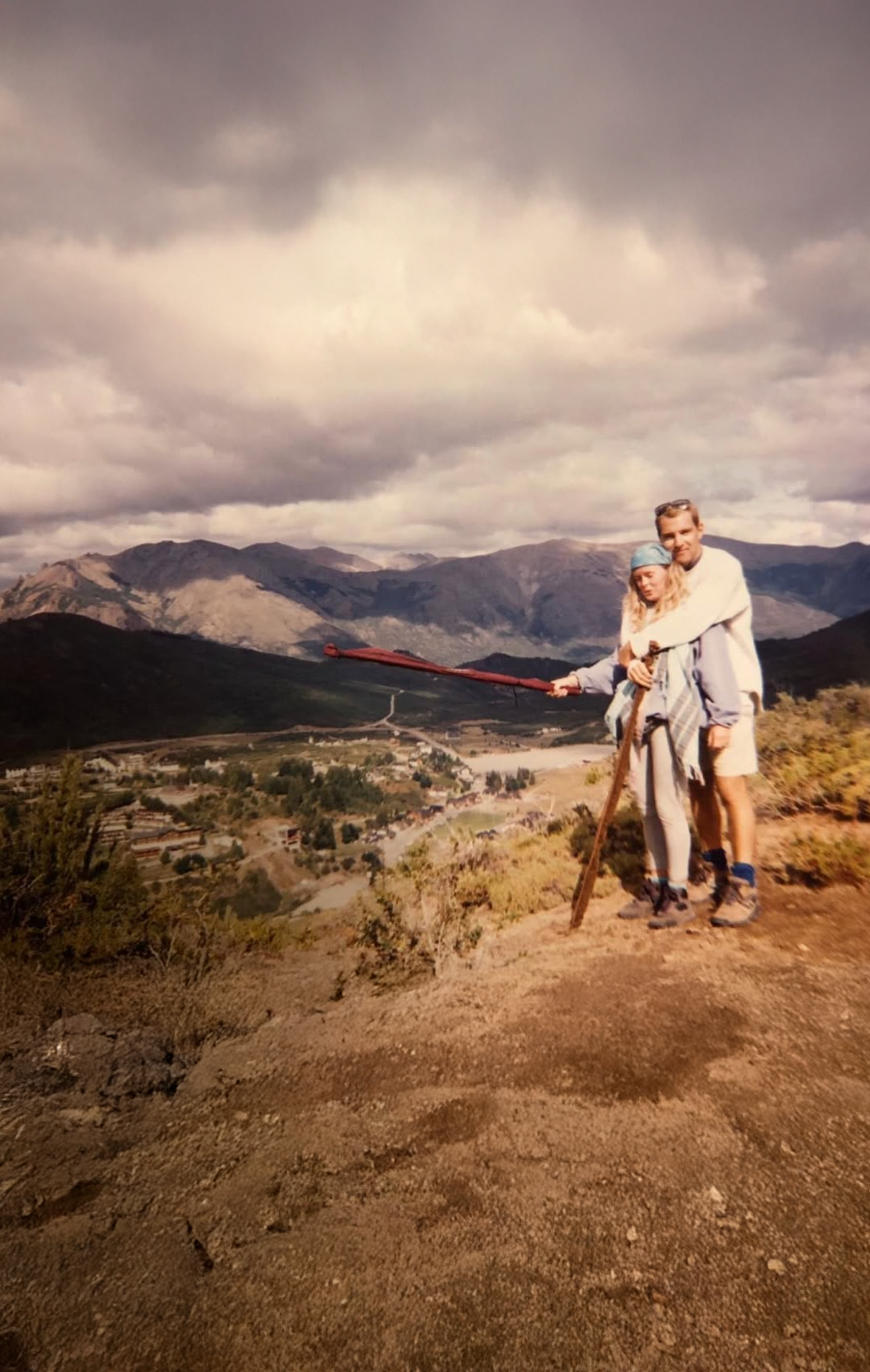 A couple standing on a hiking trail overlooking a valley and mountain range. The woman is wearing a headscarf and the man is hugging her from behind. They are both dressed in outdoor clothing and hiking boots, with a cloudy sky overhead.