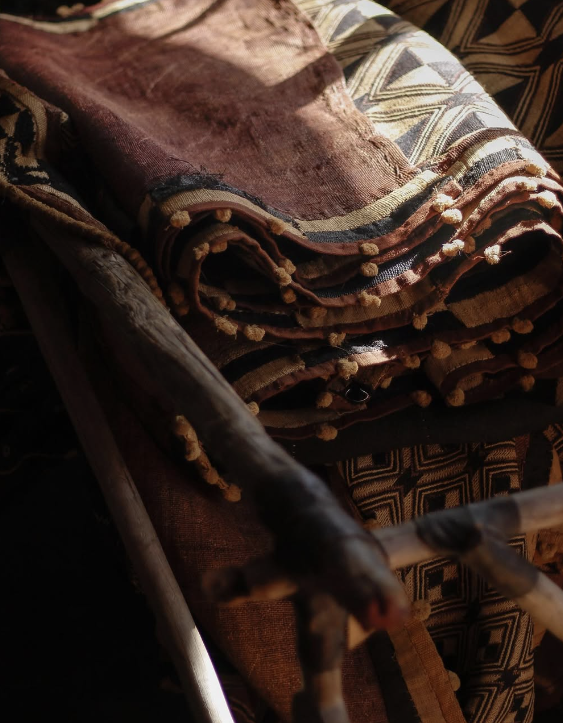 Close-up of a stack of folded patterned textiles with tassels, resting on a wooden chair with a dark background.