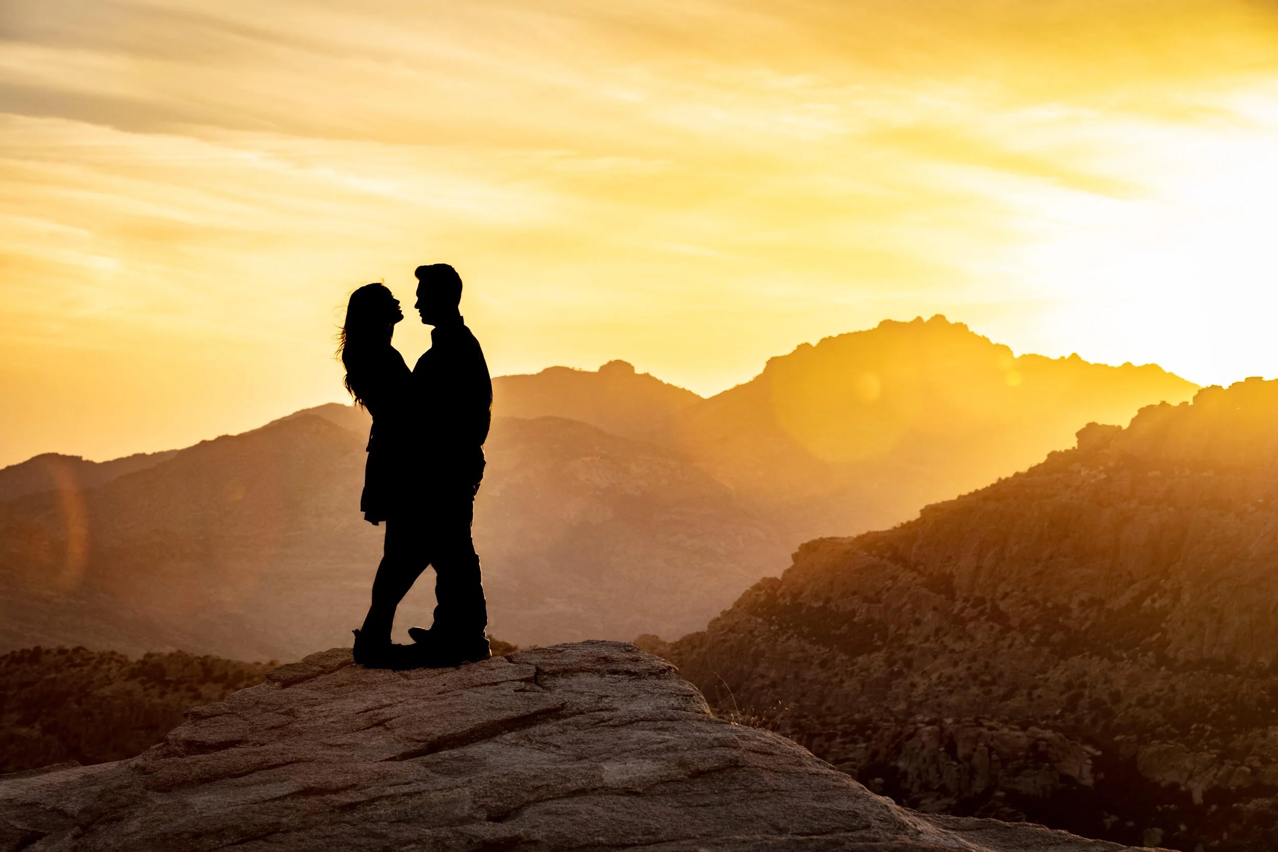 Silhouette of a couple standing on a rocky outcrop, facing each other, with a mountainous landscape and a golden sunset sky in the background.
