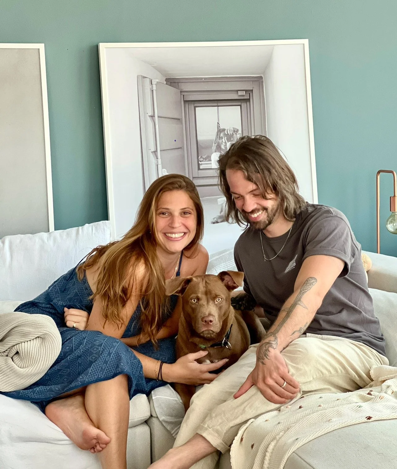 A smiling woman and man sitting on a couch with a brown dog, in a cozy living room with teal wall and wall art.