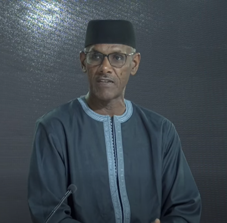 A man wearing glasses, a black hat, and a traditional blue garment standing behind a microphone against a dark background.