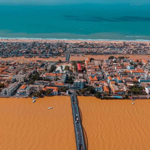 Aerial view of a coastal city showing a beach, ocean, city streets, and a bridge over a yellow-orange desert or sand dunes.
