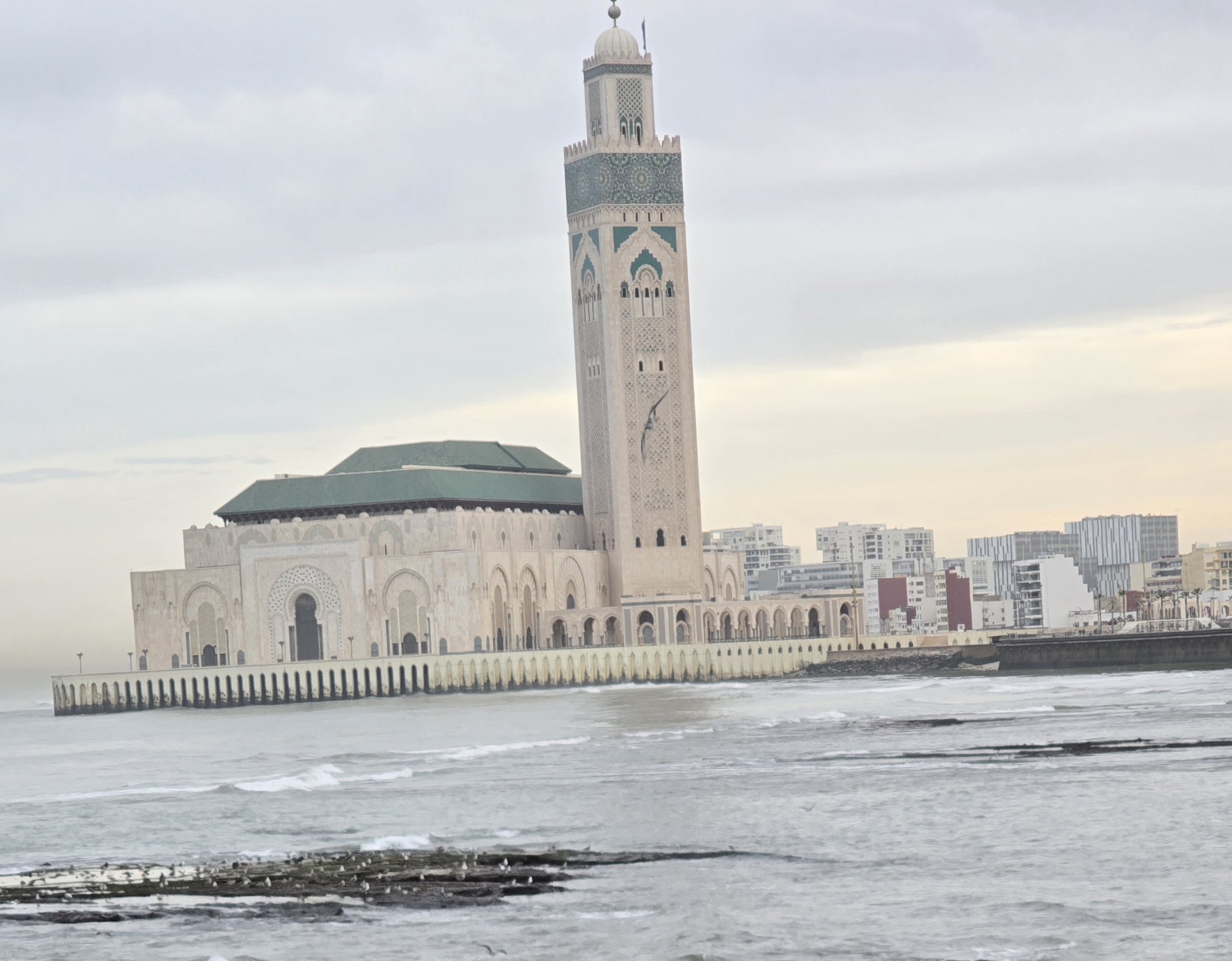 The Hassan II Mosque on the shoreline with the ocean in the foreground and modern buildings in the background.