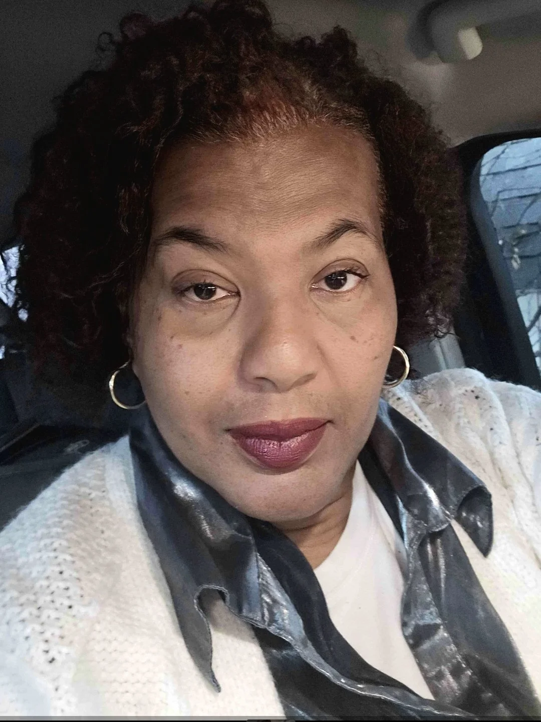 Close-up of a woman with curly hair and hoop earrings, wearing a white sweater with a black and gray satin shirt underneath, sitting in a vehicle.
