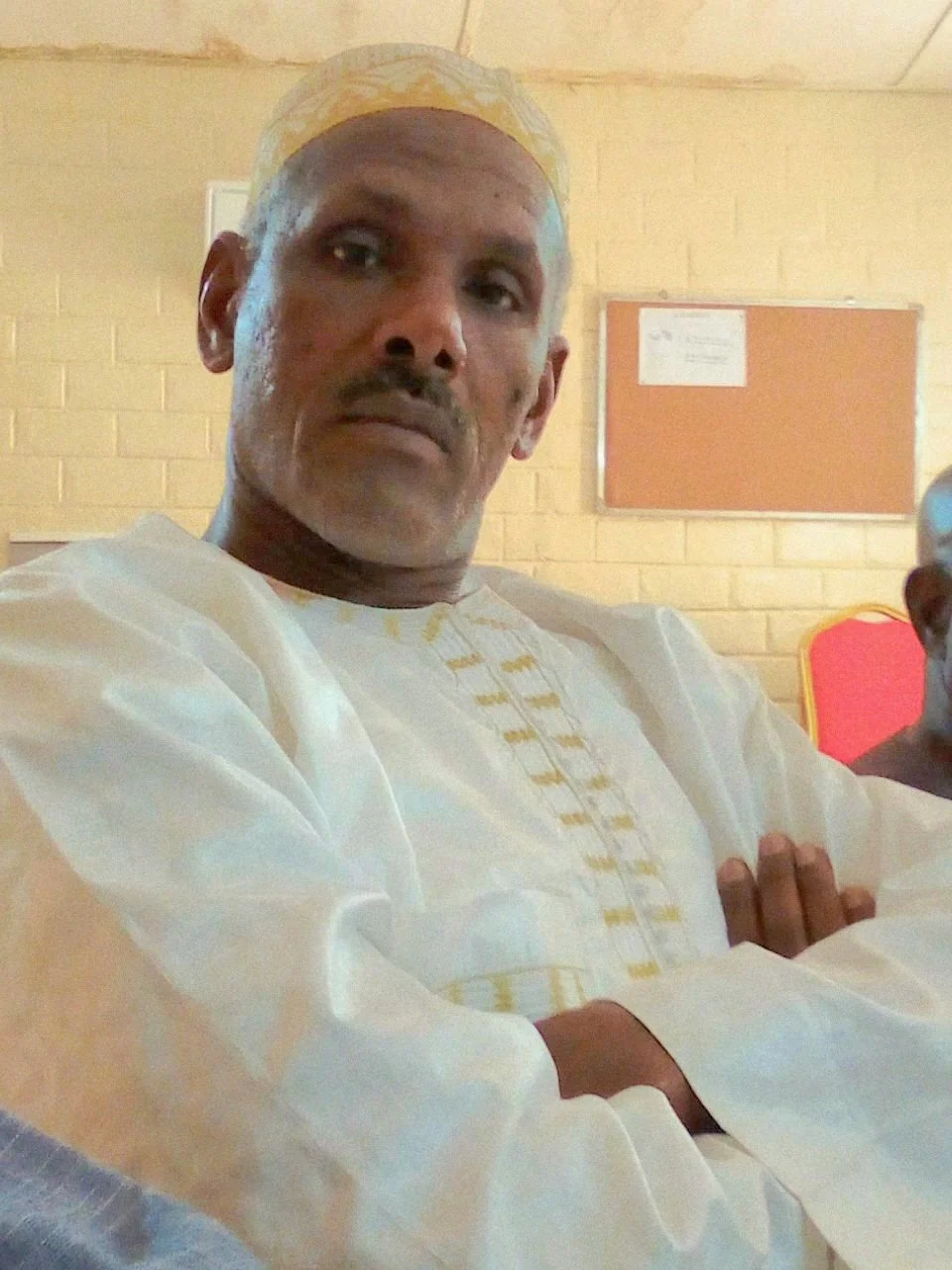 A man with crossed arms wearing a traditional white and gold outfit and a matching cap, standing indoors against a brick wall with a cork bulletin board in the background.