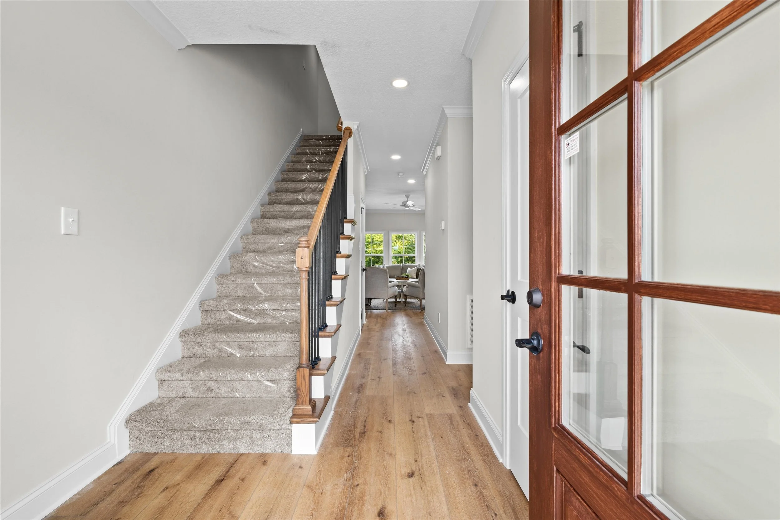 Interior view of a home entryway with a wooden front door and glass panes, a staircase with carpeted steps and wooden handrail on the left, and a hallway leading to a living area with large windows and a ceiling fan.