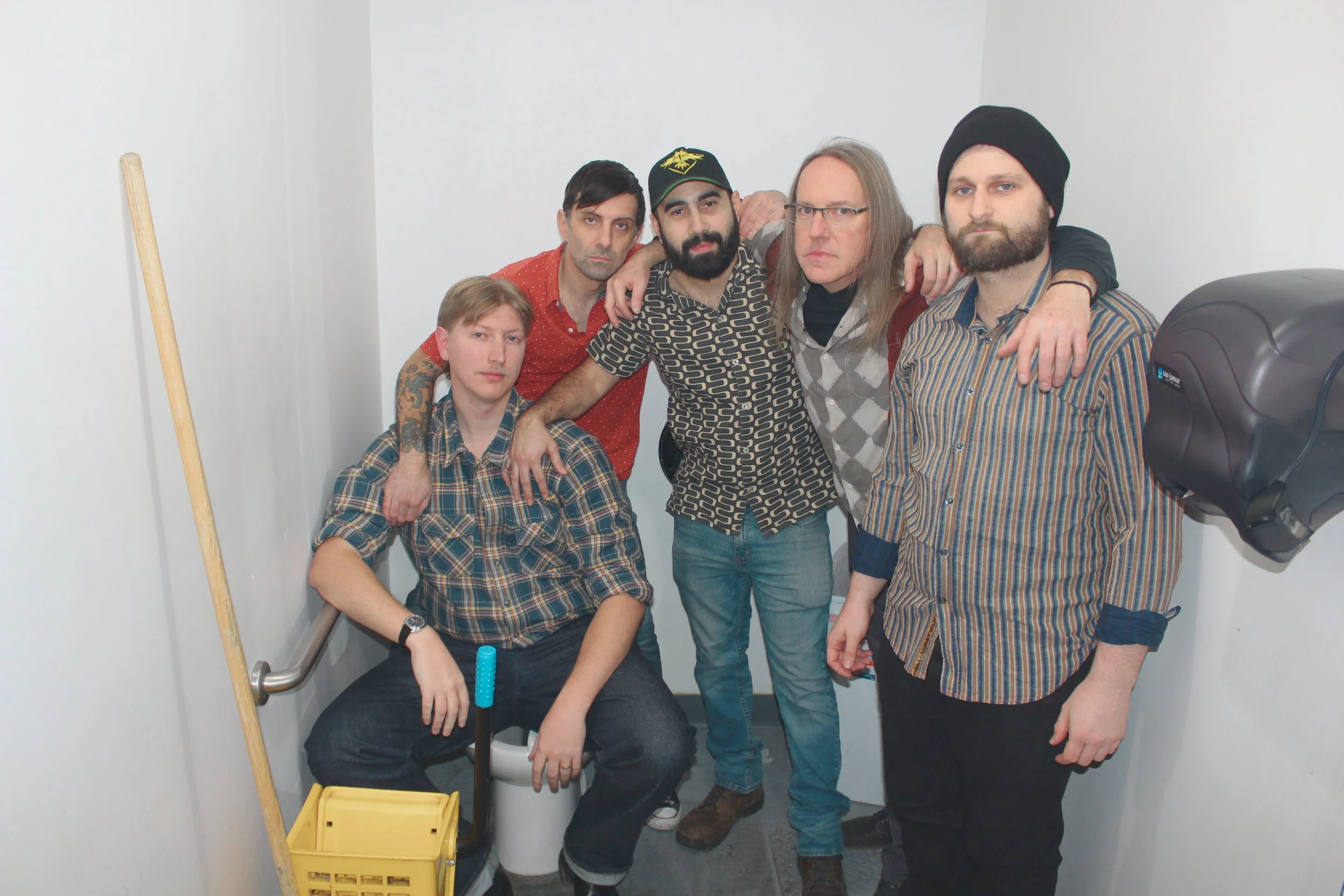 Five men in a small, white bathroom posing for a photo. One man is sitting on a toilet with a yellow cleaning bucket and mop nearby, while the other four are standing around him. The group is dressed casually in patterned shirts and jeans, with some wearing hats or glasses.