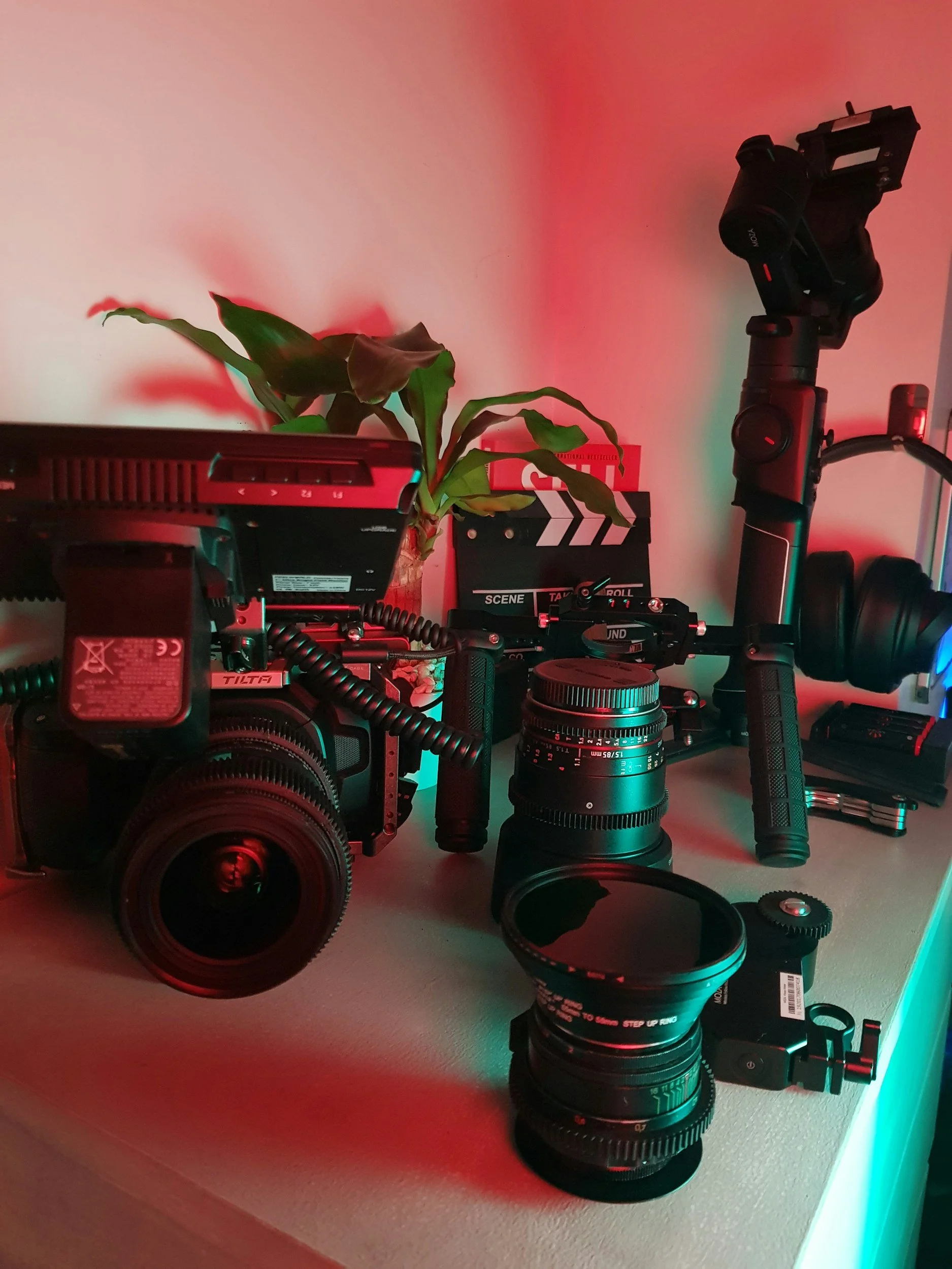 Camera equipment, lenses, headphones, and a potted plant with a clapperboard in the background on a desk.