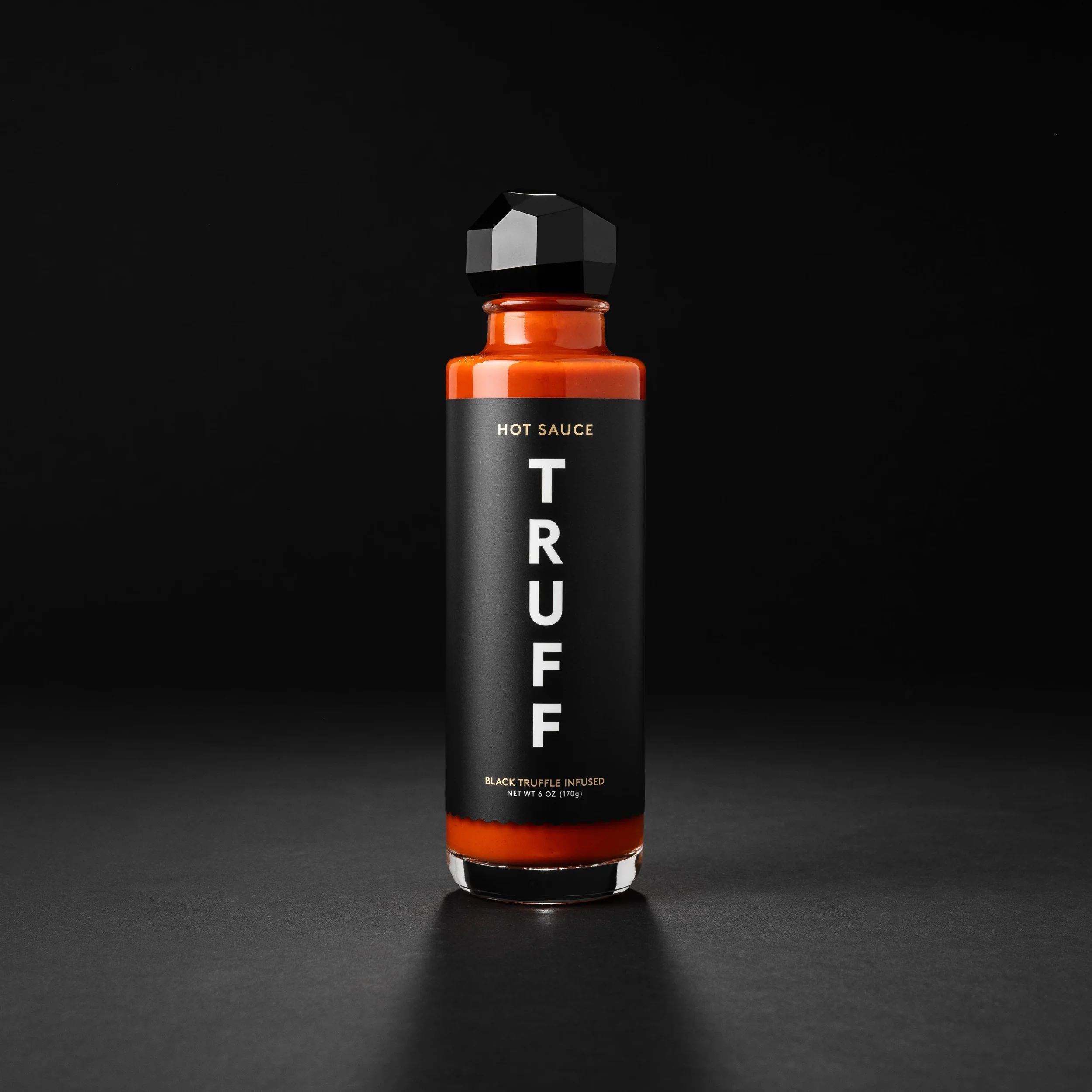 A black bottle labeled "TRUFF" with a black geometric cap, containing hot sauce infused with black truffle, against a black background.