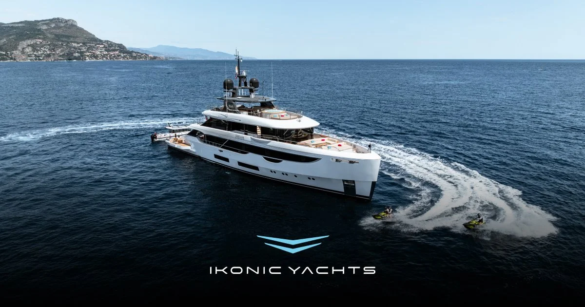 A large white yacht sailing on the ocean near a coastal landmass with hills, under a clear blue sky, with two people riding jet skis nearby, and the Ikonic Yachts logo at the bottom.