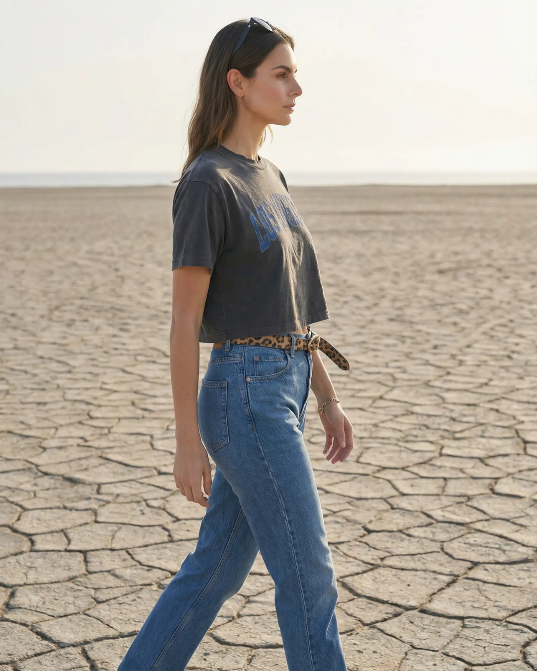 Young woman walking in a dry, cracked landscape with a flat horizon, wearing sunglasses, a black crop top, blue jeans, and a leopard print belt.