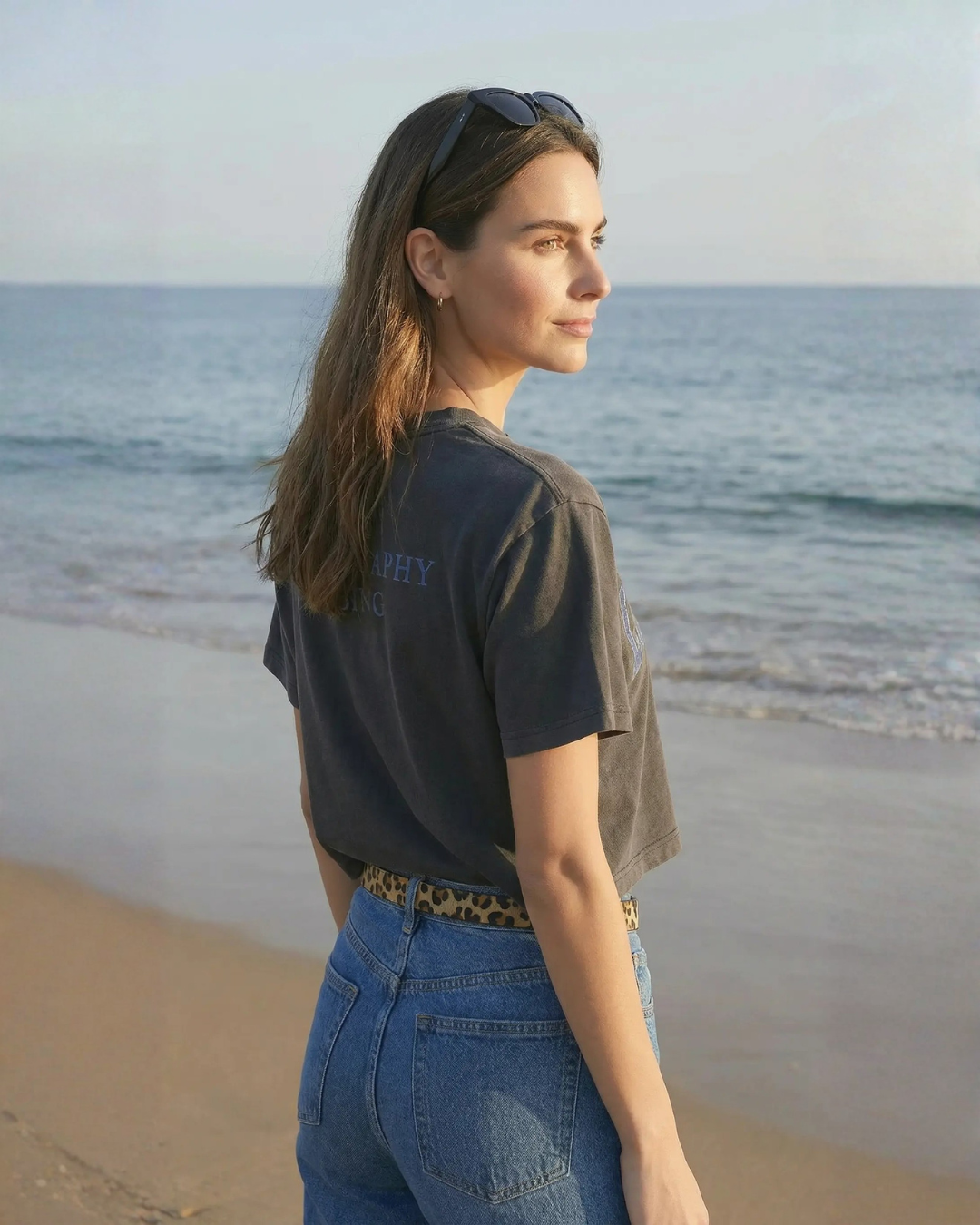 A woman with long brown hair standing on the beach, looking out at the ocean during sunset, wearing sunglasses on her head, a black T-shirt, and blue jeans.
